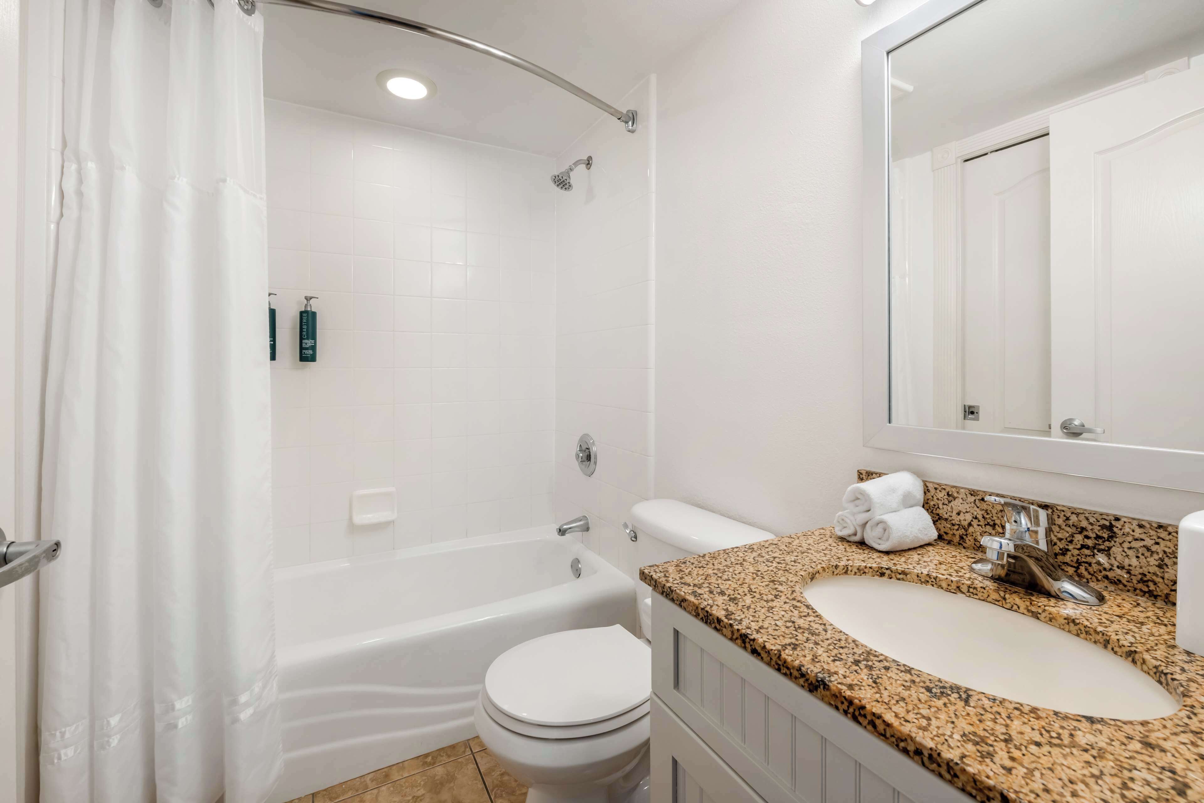 Une salle de bain avec une baignoire blanche, des toilettes et un lavabo, le tout surmonté d’un plan de travail en granit.