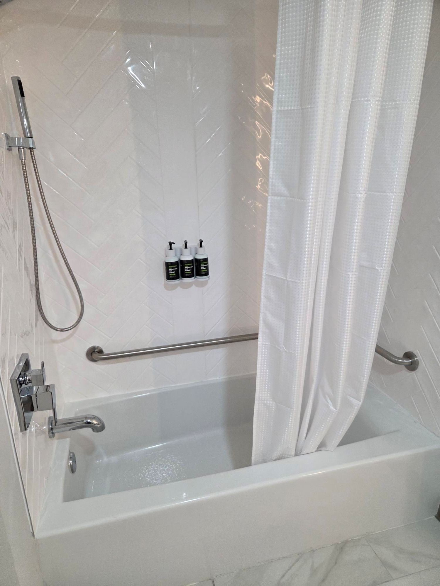 A white bathtub with a showerhead and handrails, a towel rack, and three bottles of shampoo.