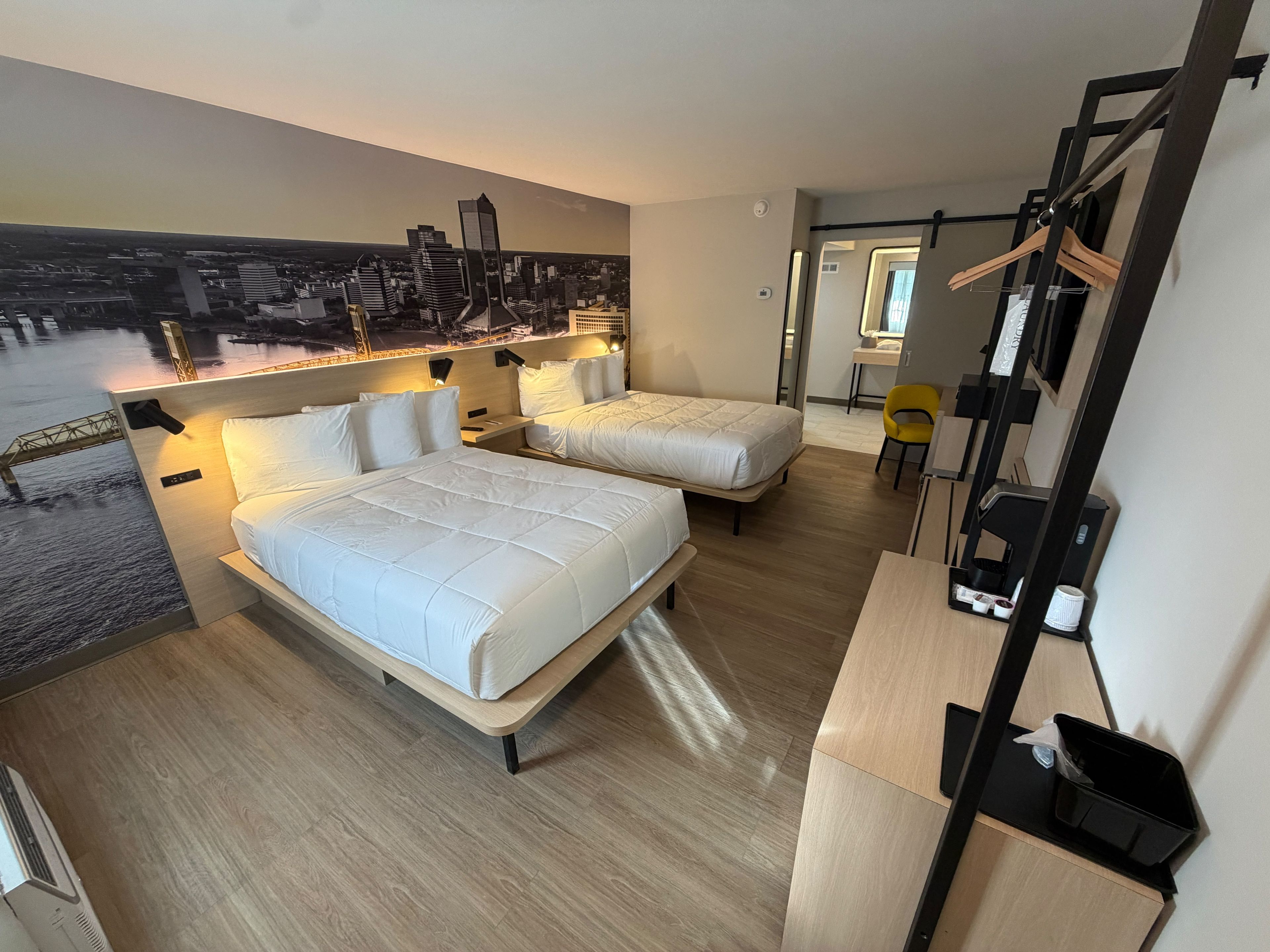 A hotel room with two beds, a cityscape mural, a desk, and a chair.