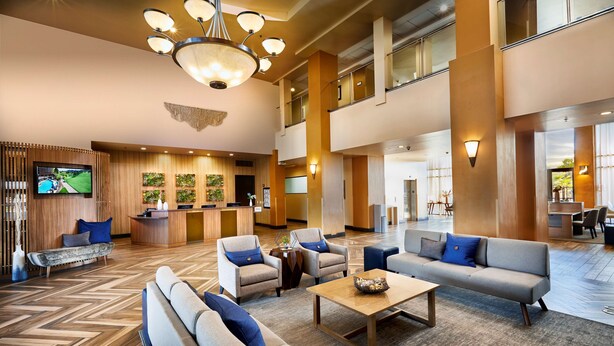 Eine großzügige Lobby mit hoher Decke, mehreren Sitzbereichen, einem Empfangsbereich und einem Fernsehbildschirm.