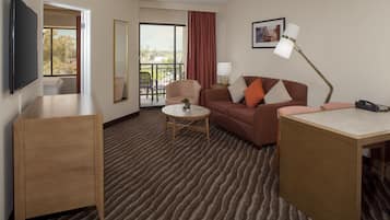 Habitación de hotel con sofá, una mesita, escritorio, televisor, lámpara y balcón con vistas.