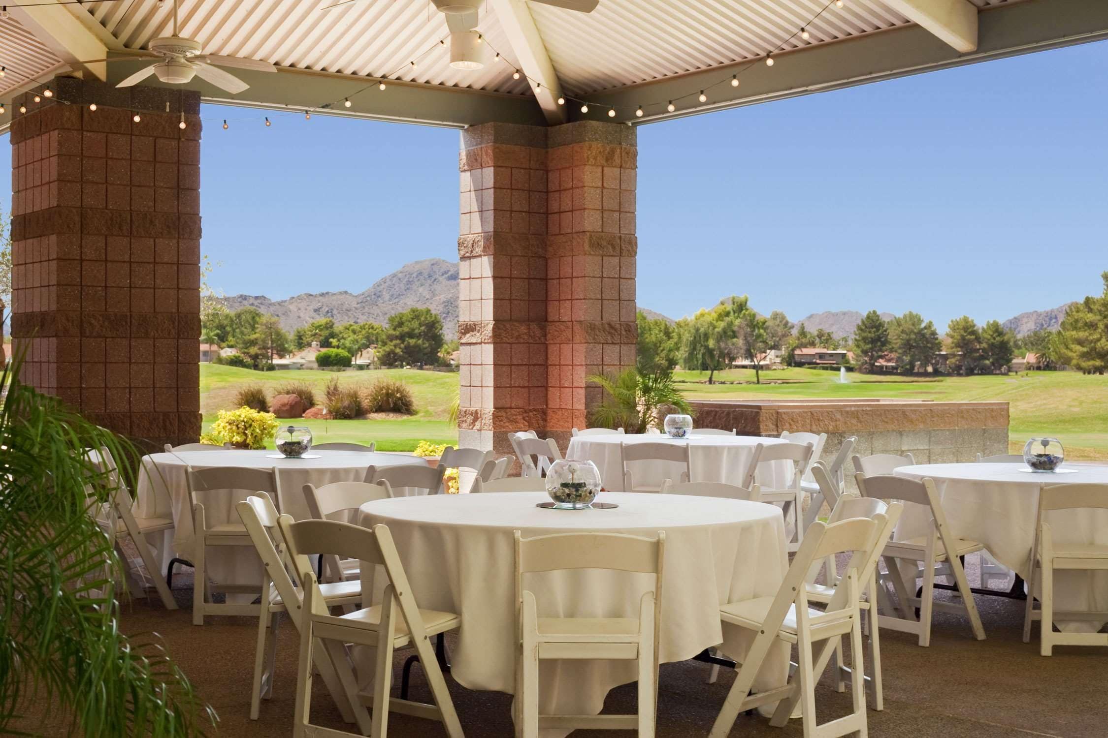An outdoor patio with round tables and white chairs, set against a backdrop of mountains and a golf course.