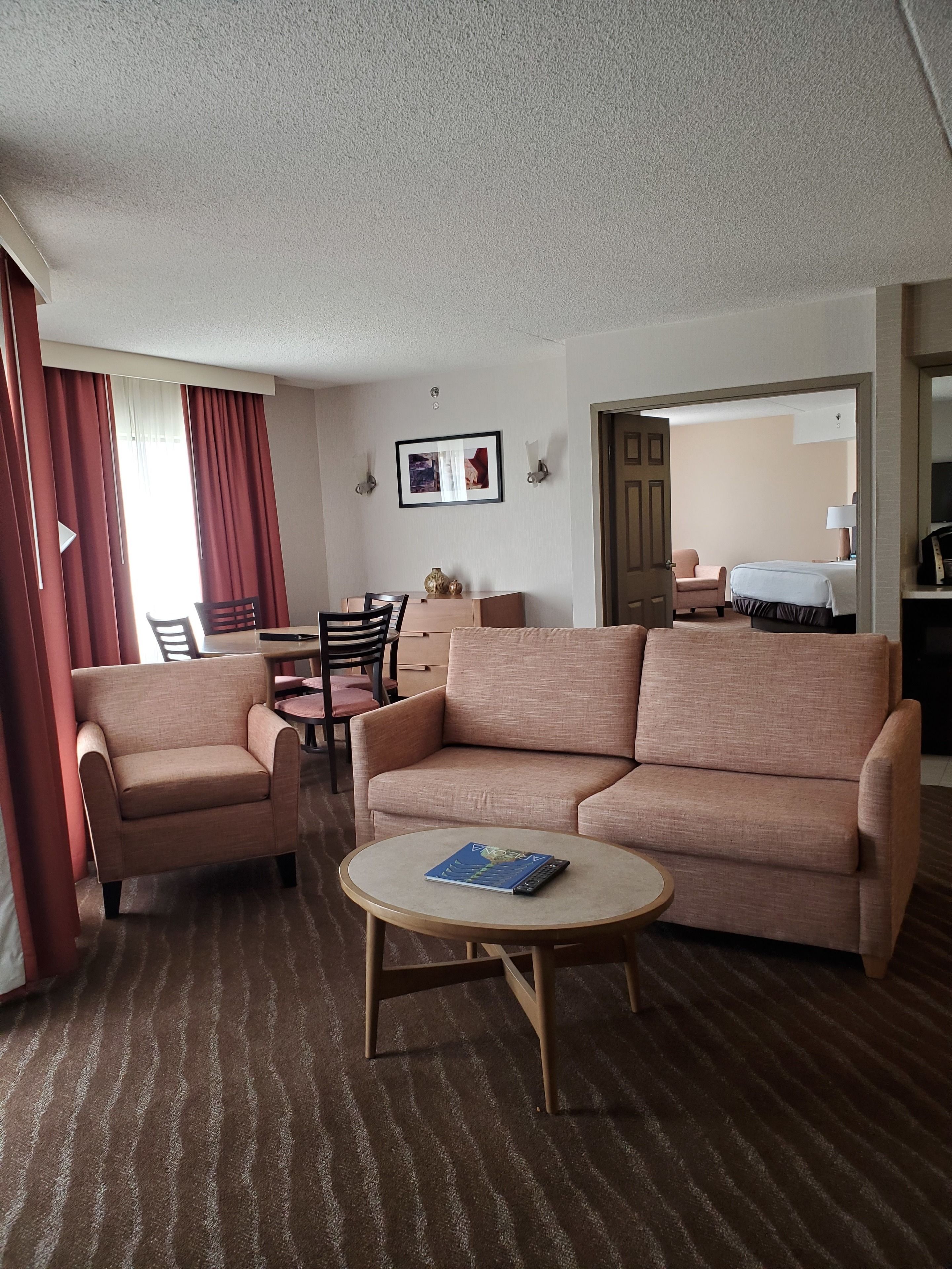 A hotel room with a sofa, armchair, round coffee table, and a dining area with chairs and a table.