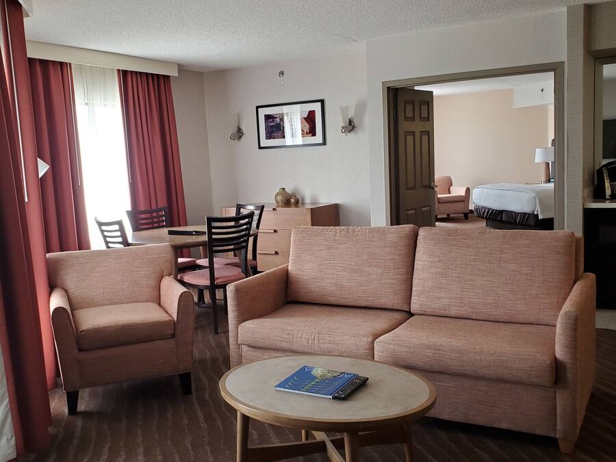 A hotel room with a sofa, armchair, round coffee table, and a dining area with chairs and a table.