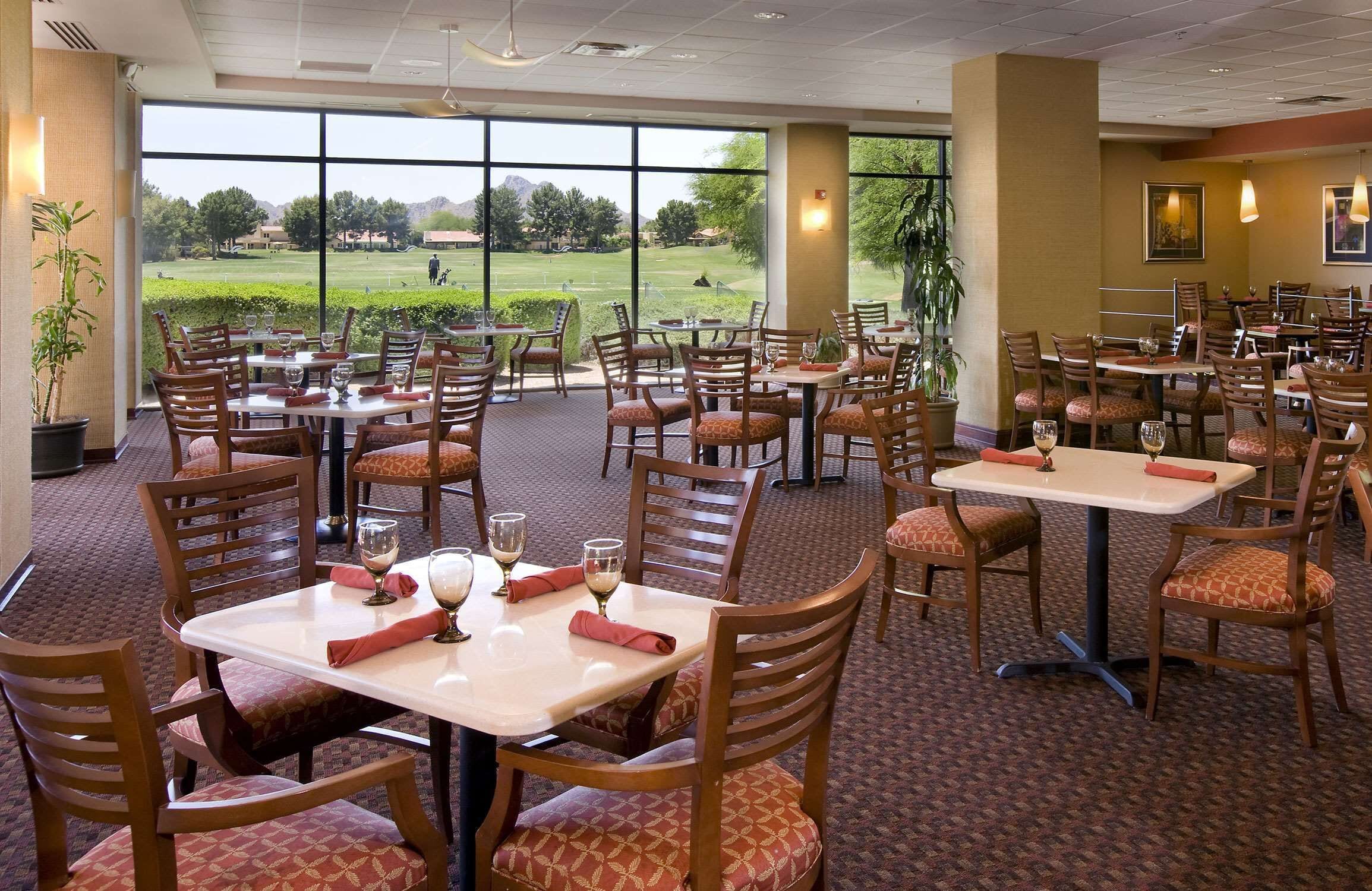 A restaurant with multiple tables and chairs, large windows offering a view of a golf course, and indoor lighting.