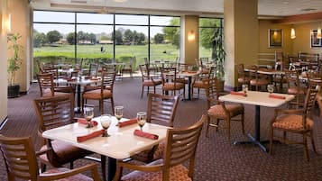 A restaurant with multiple tables and chairs, large windows offering a view of a golf course, and indoor lighting.