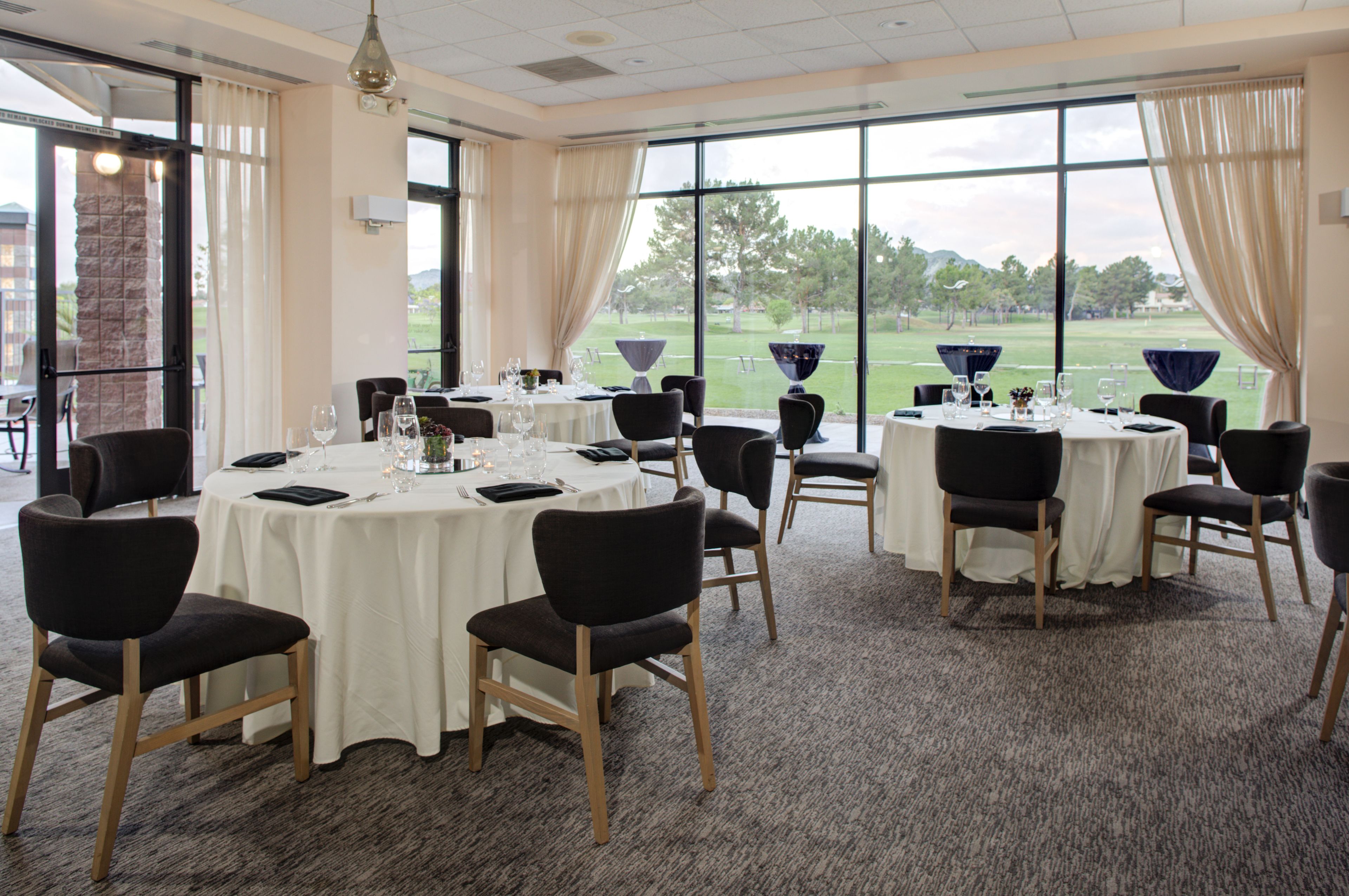 A conference room with round tables and chairs, large windows with curtains, and a view of a golf course.