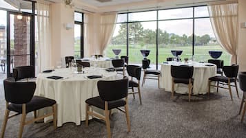 A conference room with round tables and chairs, large windows with curtains, and a view of a golf course.