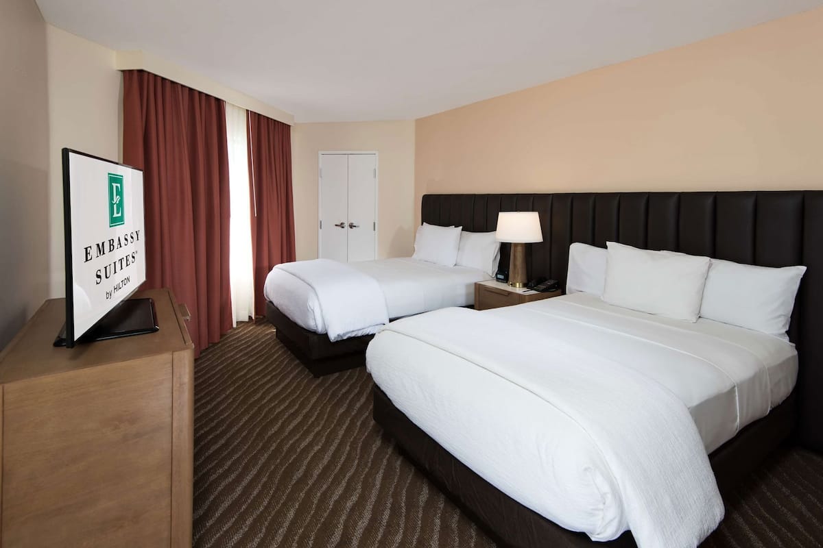 Quarto de hotel com duas camas, uma escrivaninha, uma luminária e uma placa indicando ser um Embassy Suites by Hilton.