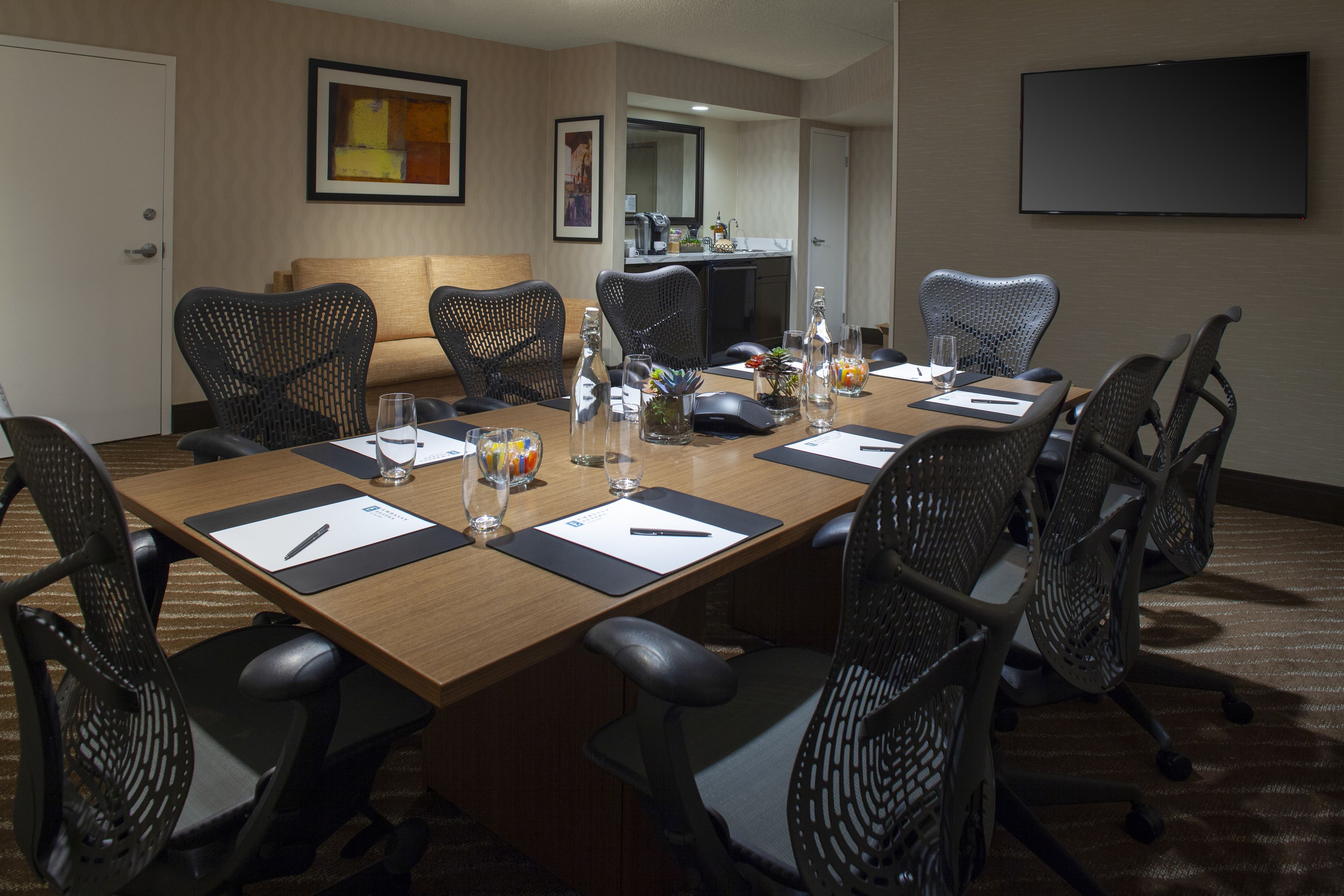 A conference room with a long table, black mesh chairs, a sofa, a TV, and a kitchen area in the background.