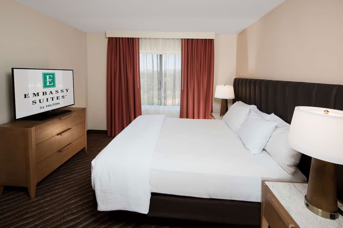 Quarto de hotel com cama, televisão, escrivaninha, luminária e janela com cortinas.