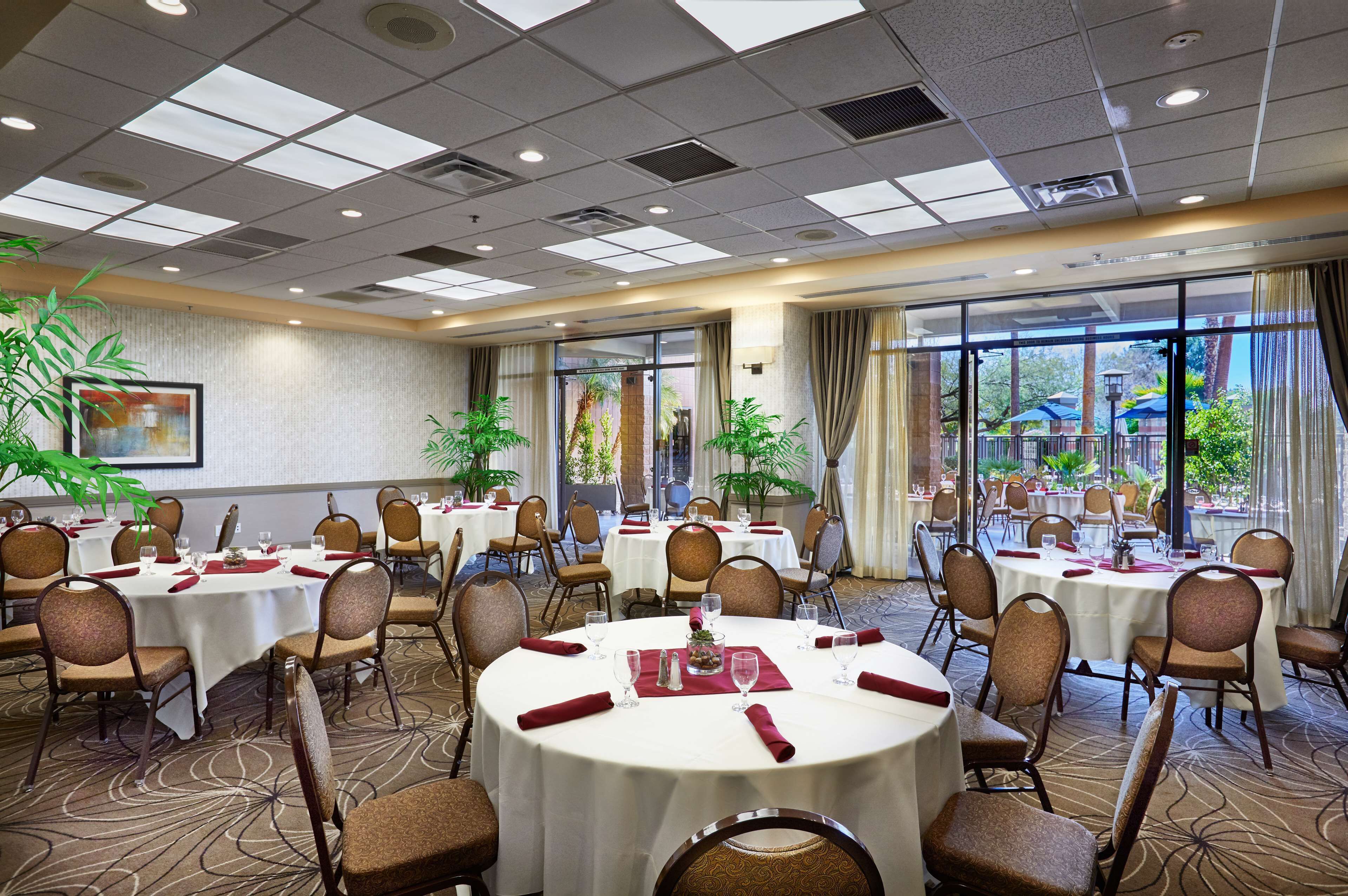 A hotel dining area with round tables, chairs, and large windows offering a view of the outdoors.