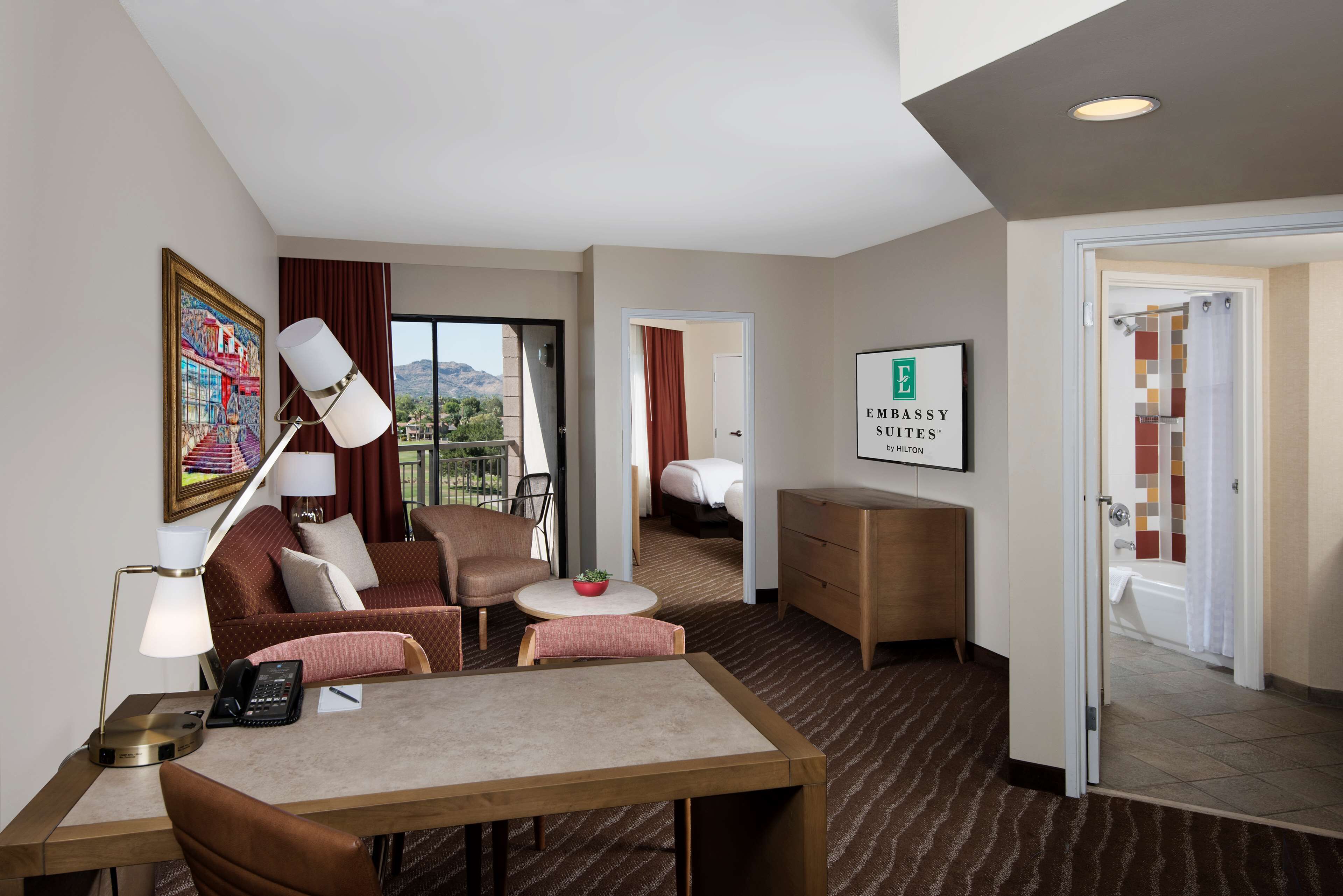 Ein Hotelzimmer mit Schreibtisch, Sofa, Sesseln, einem kleinen Tisch mit Pflanze, einer Lampe, einem Bett und einem durch eine geöffnete Tür einsehbaren Badezimmer.