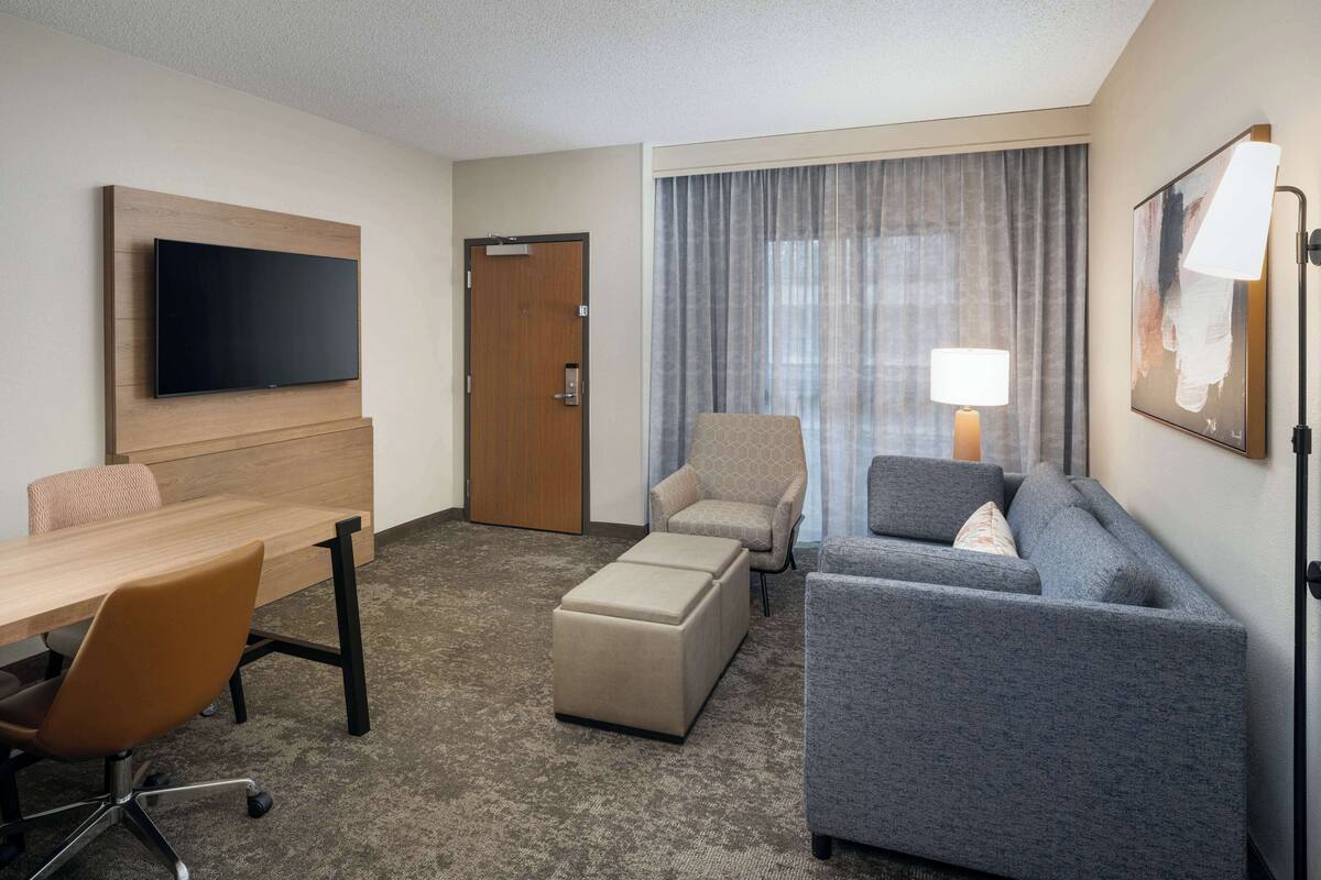 A hotel room with a flat-screen TV, a wooden desk, a grey sofa, a beige ottoman, a patterned armchair, and a large window with curtains.