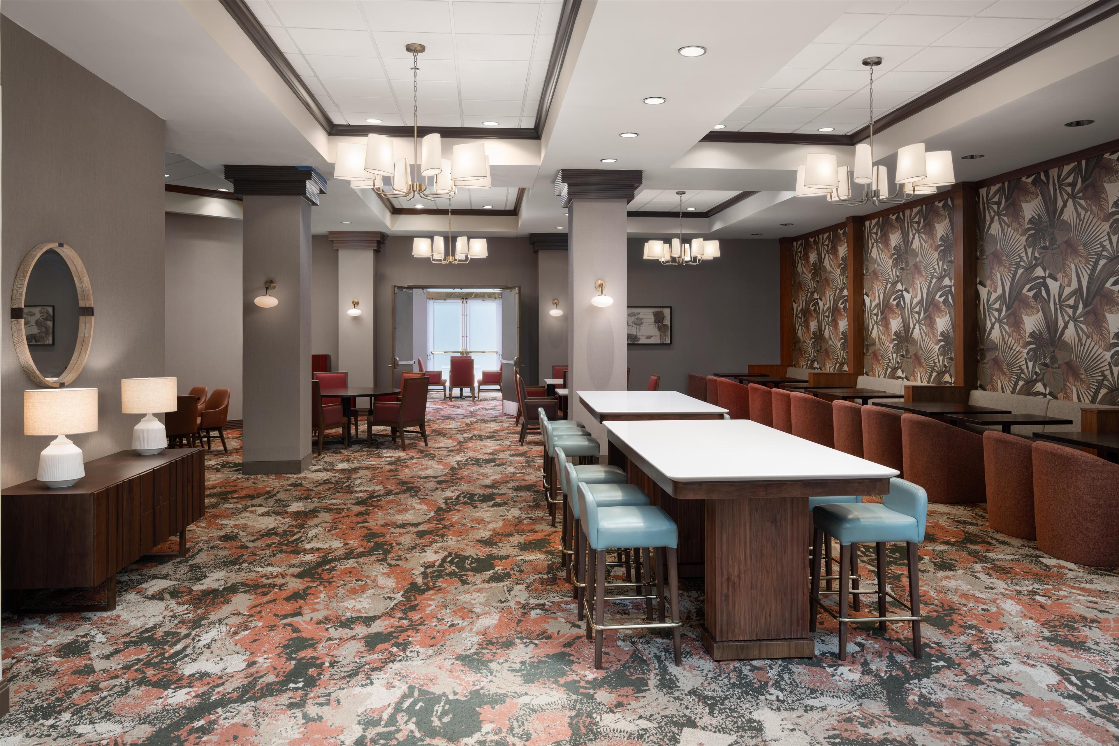 A modern hotel lobby with a patterned carpet, bar area with stools, and a large window in the background.