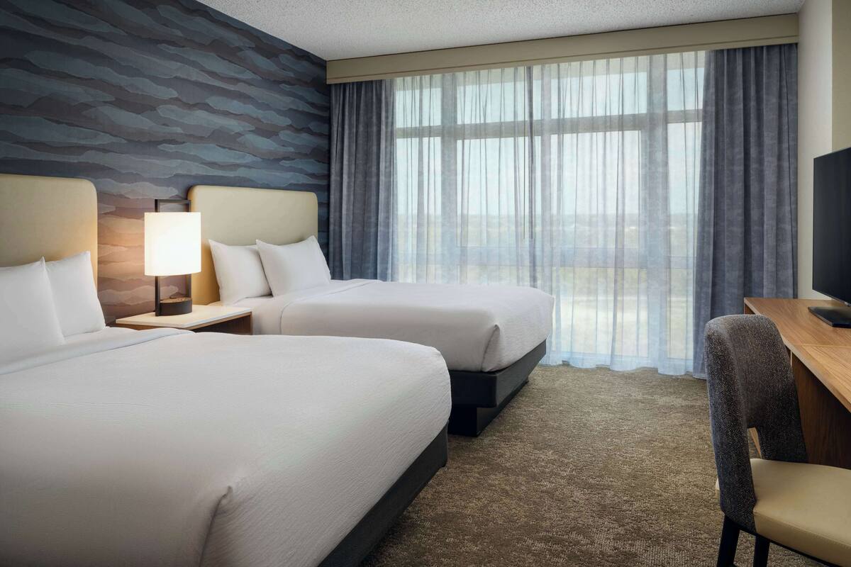 A hotel room with two beds, a desk, a chair, a lamp, and a large window with curtains.