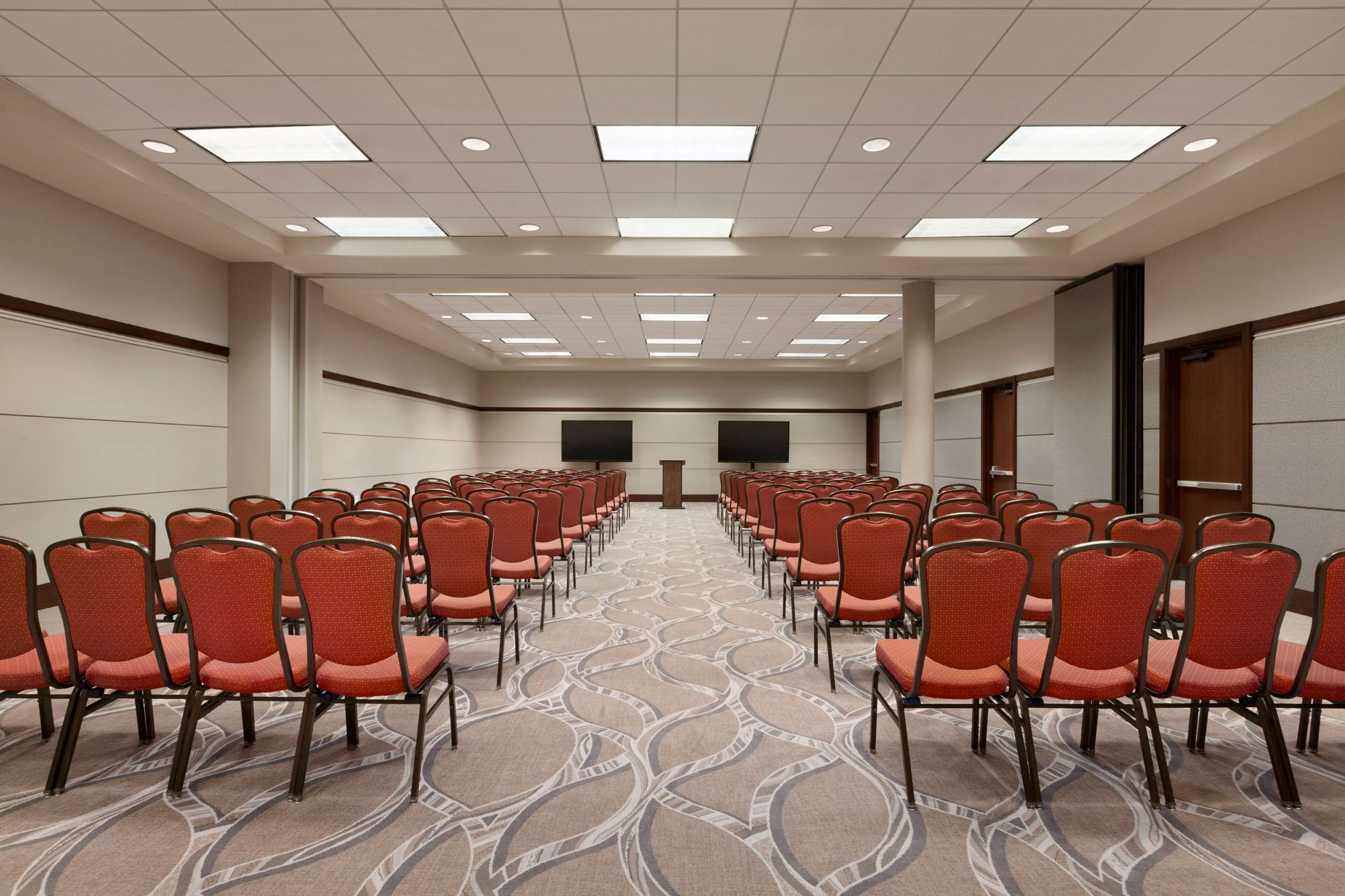 Sala de conferências com fileiras de cadeiras laranja, carpete estampado e teto com iluminação embutida.