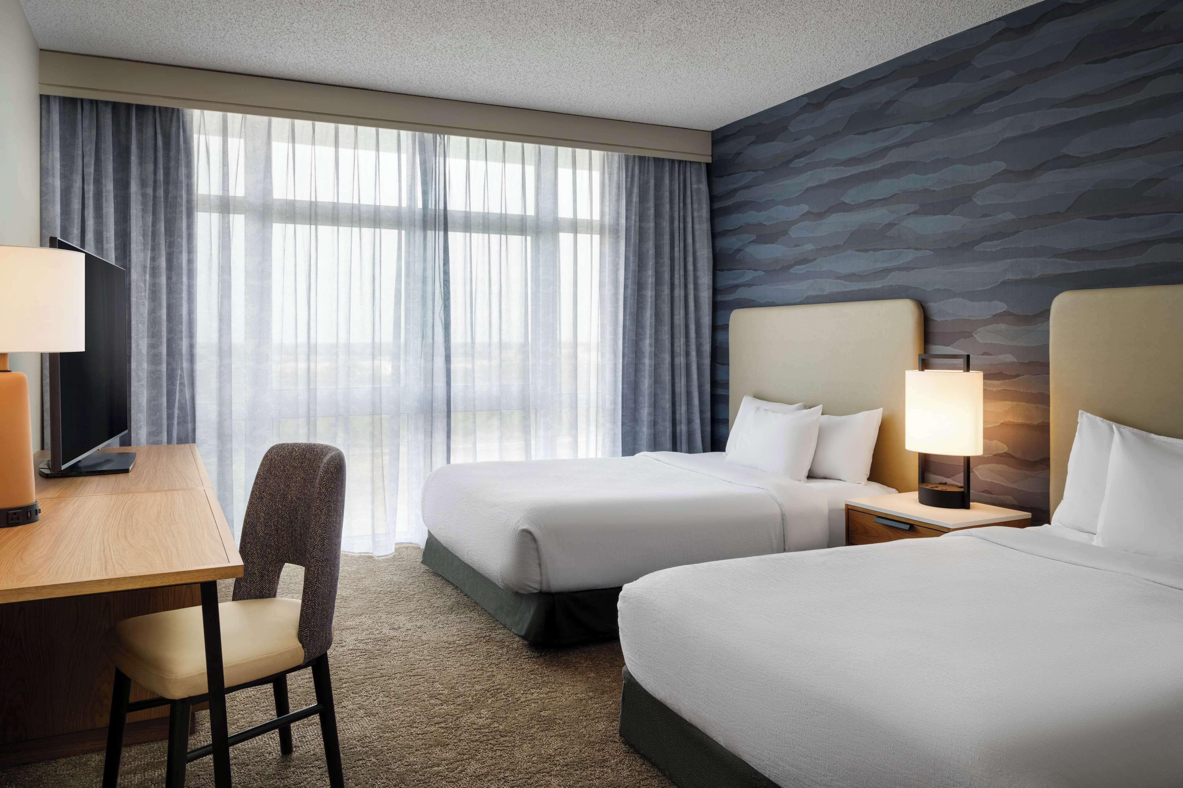 A hotel room with two beds, a desk with a chair, a TV, and a window with sheer curtains.