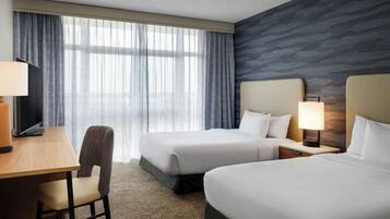 A hotel room with two beds, a desk with a chair, a TV, and a window with sheer curtains.