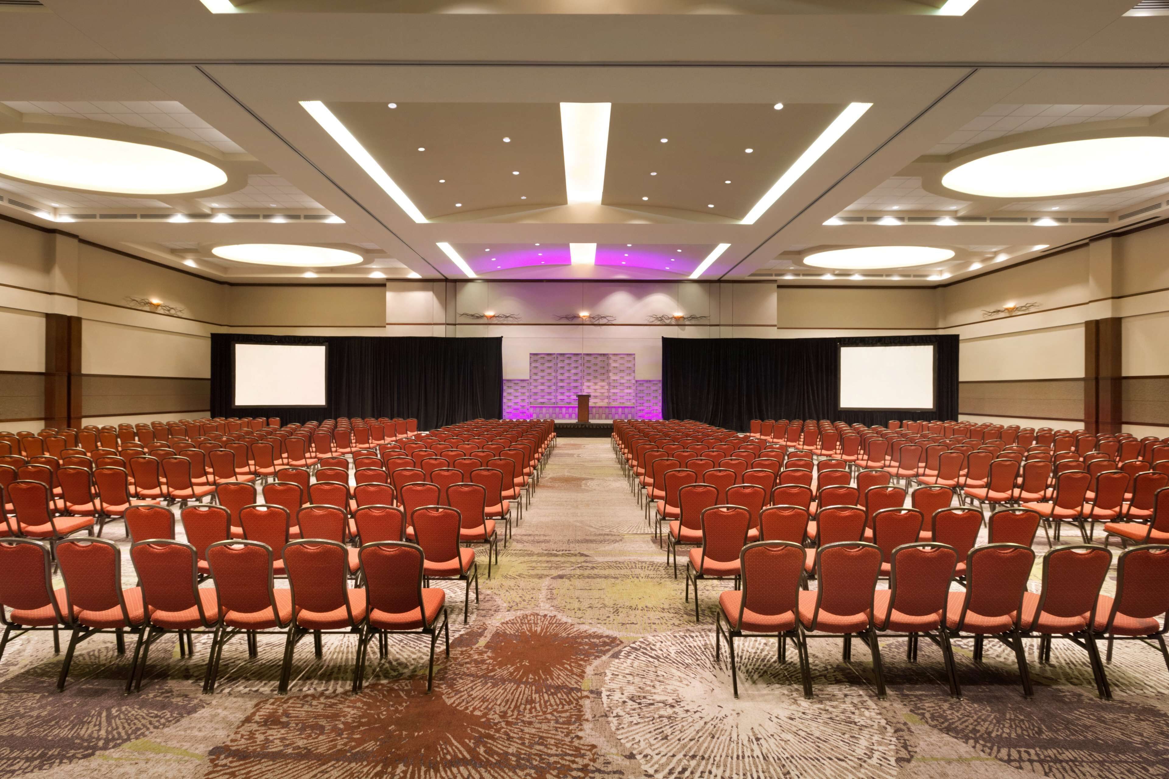 Ein Konferenzraum mit Reihen roter Stühle, einer Bühne mit violettem Hintergrund und einer großen Leinwand im Vordergrund.