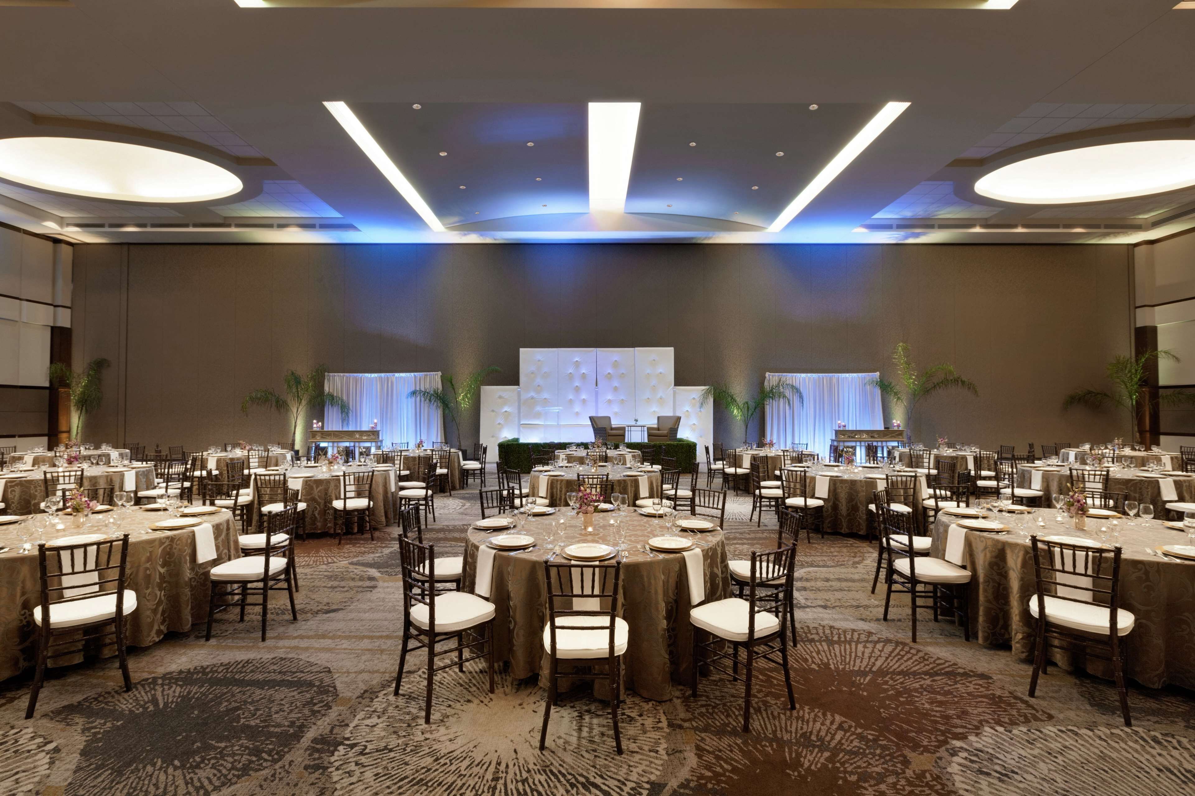 Un amplio salón de banquetes con mesas redondas dispuestas para comer, un escenario al frente y suelo alfombrado.