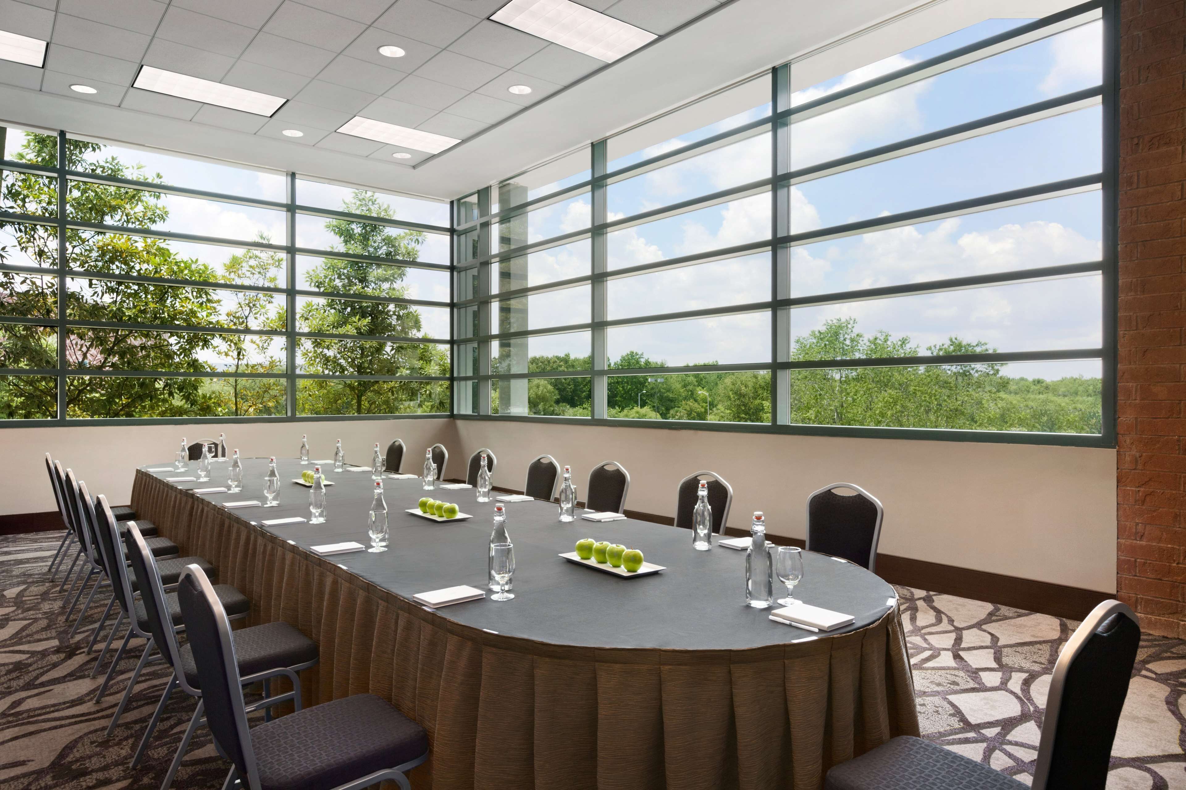 A conference room with a long table, chairs, water bottles, and a view of trees through large windows.