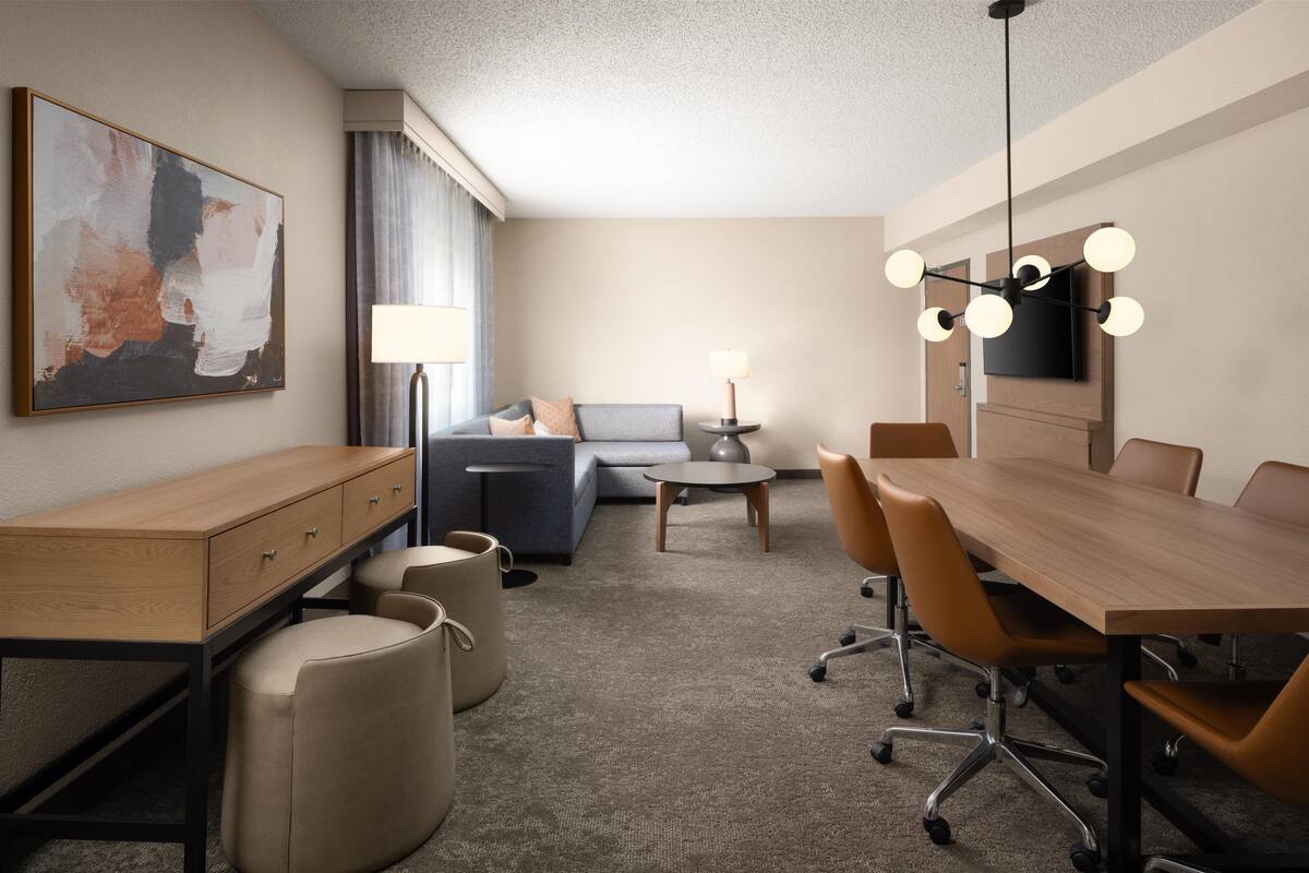 Una habitación de hotel moderna con un escritorio de madera, un sofá, una mesa de comedor con sillas, un televisor montado en la pared y una lámpara de techo.