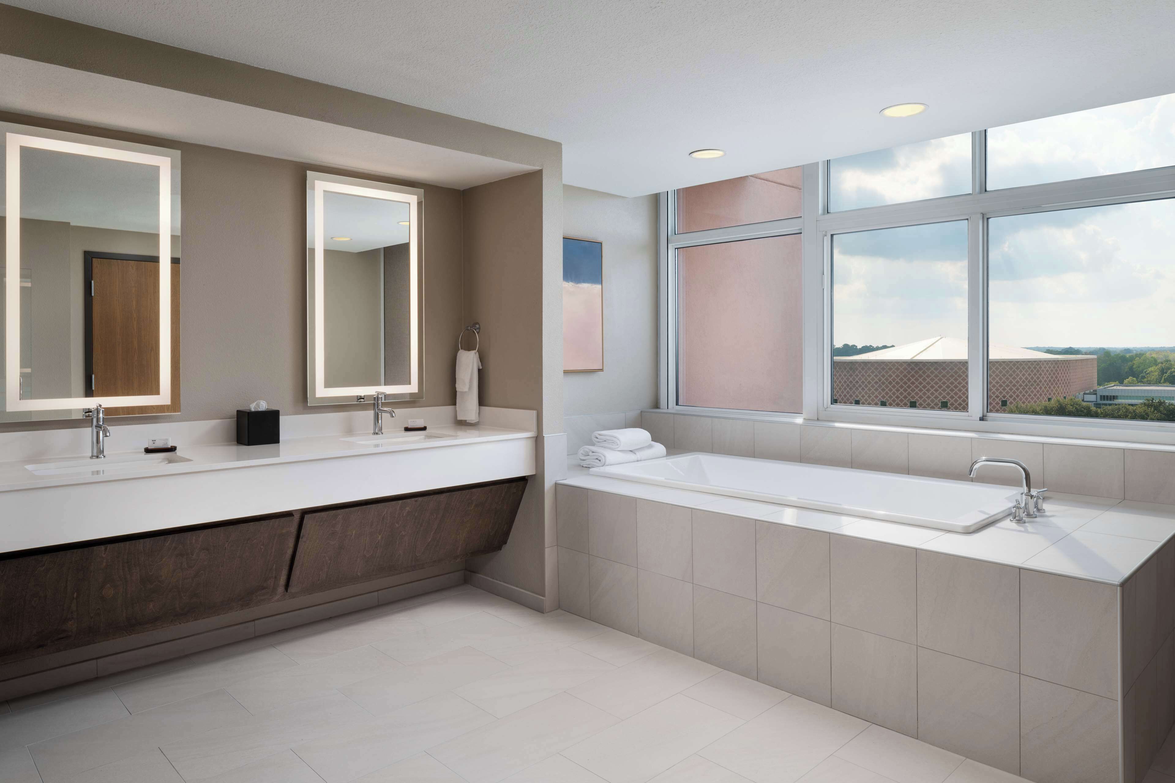A modern bathroom with two sinks, a large bathtub, and a window providing a view of the outside.