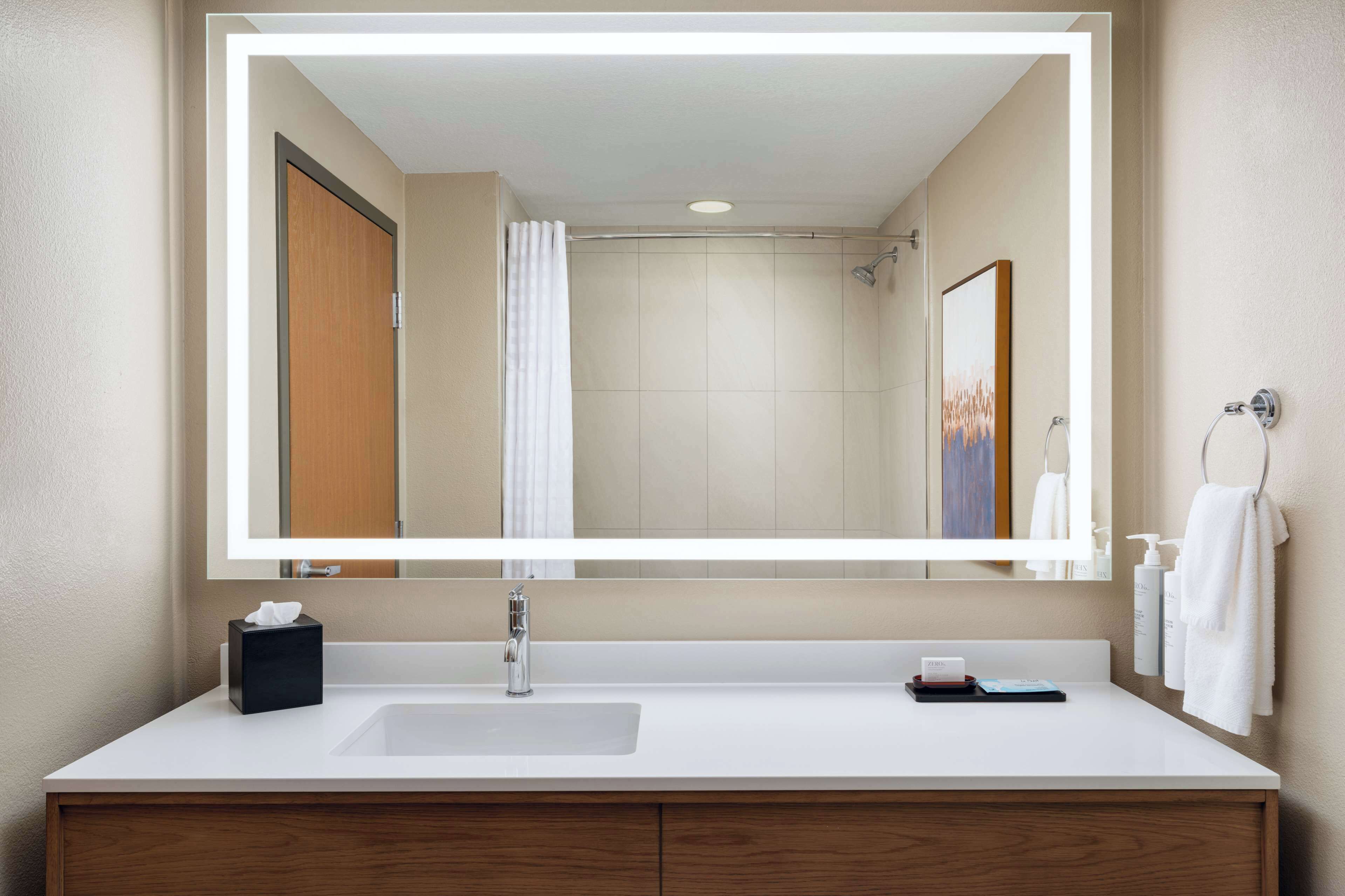A modern bathroom with a large mirror, a white sink, a wooden vanity, and a shower area.