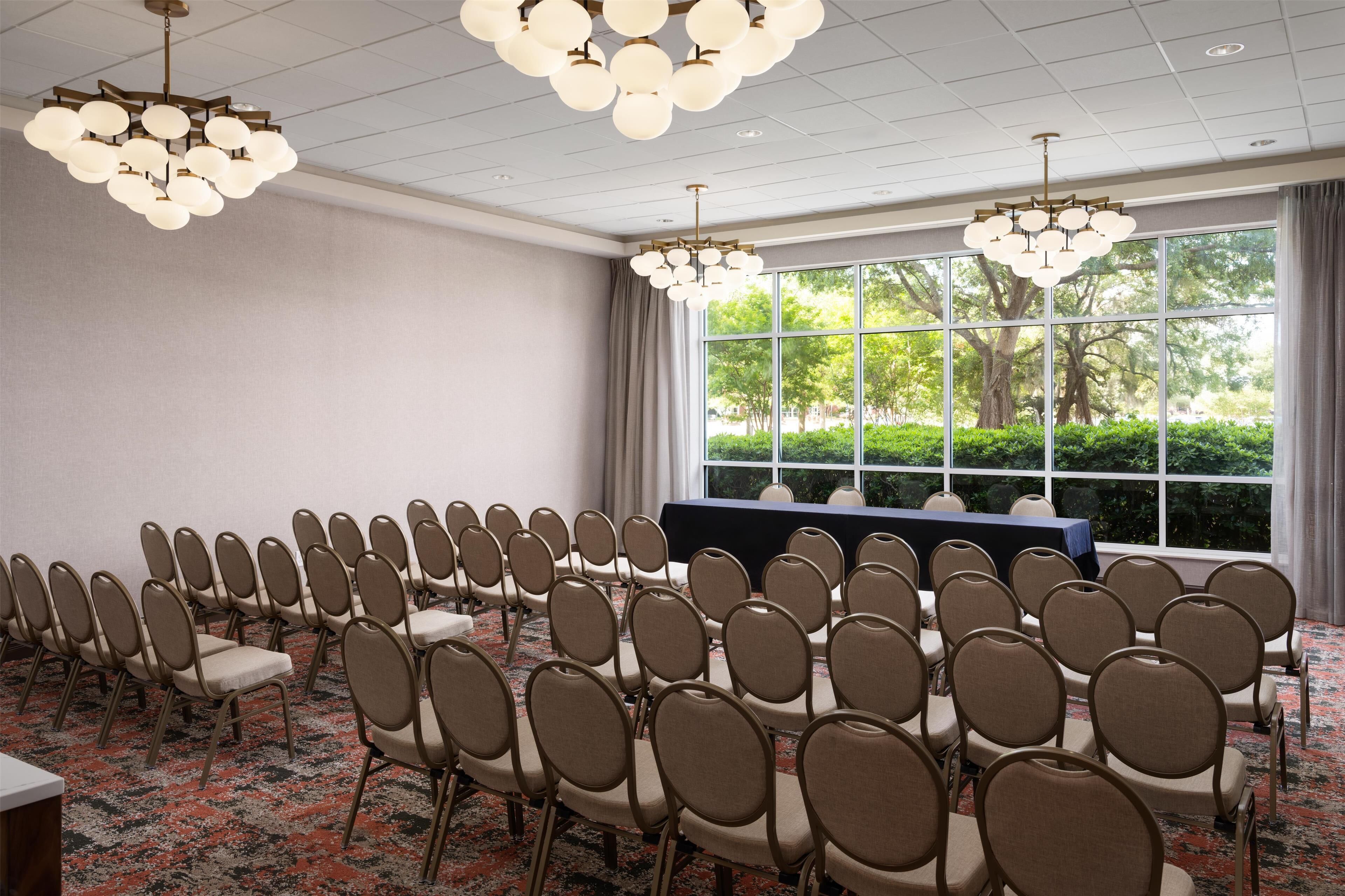 Un salón de conferencias con filas de sillas, una mesa y ventanas amplias que dan a zonas verdes.