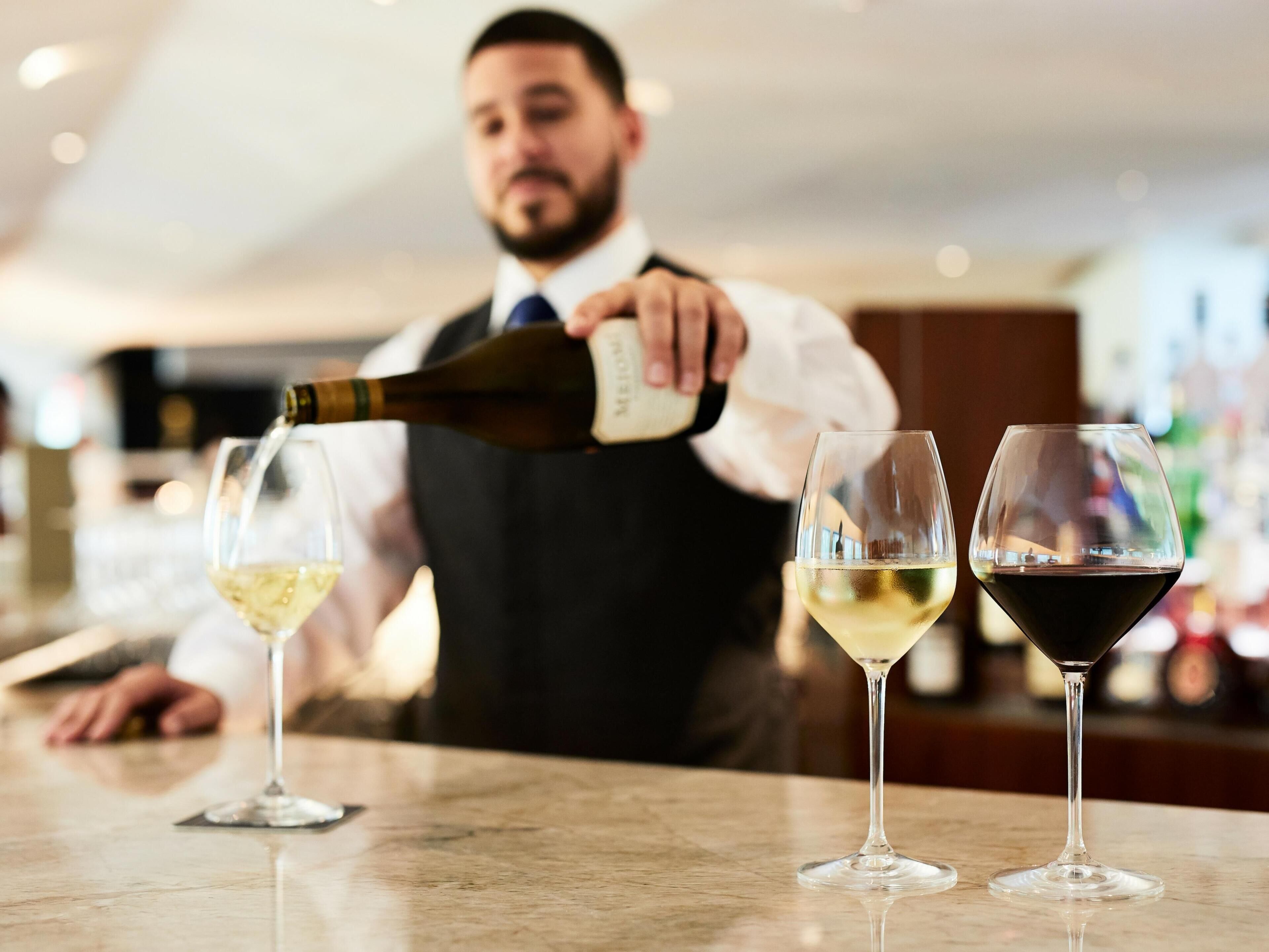 A bar setting with a person pouring wine into glasses, three glasses on the counter, and a blurred background.