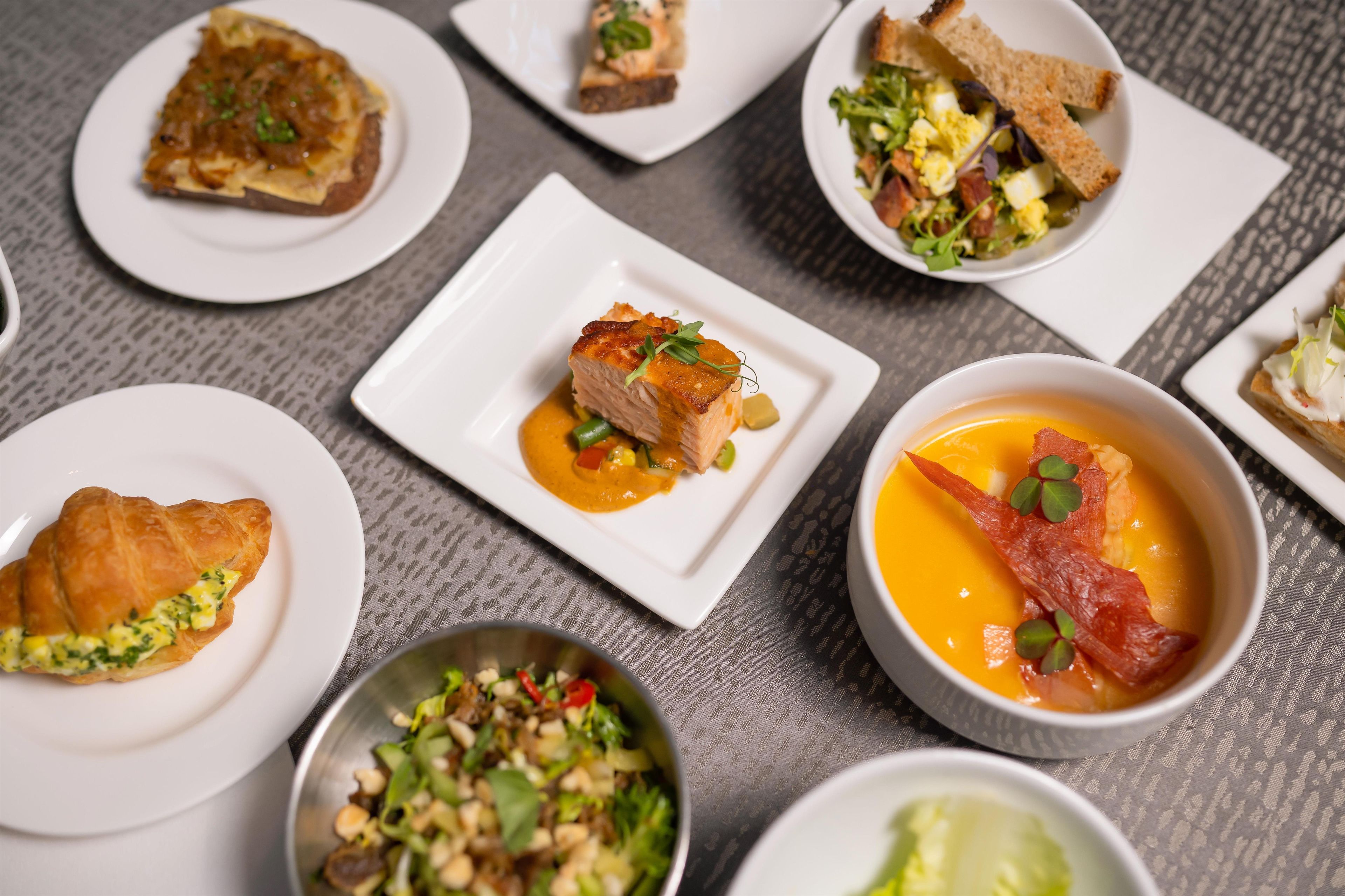 A variety of dishes on white plates, including a croissant, a bowl of soup, and a salad.
