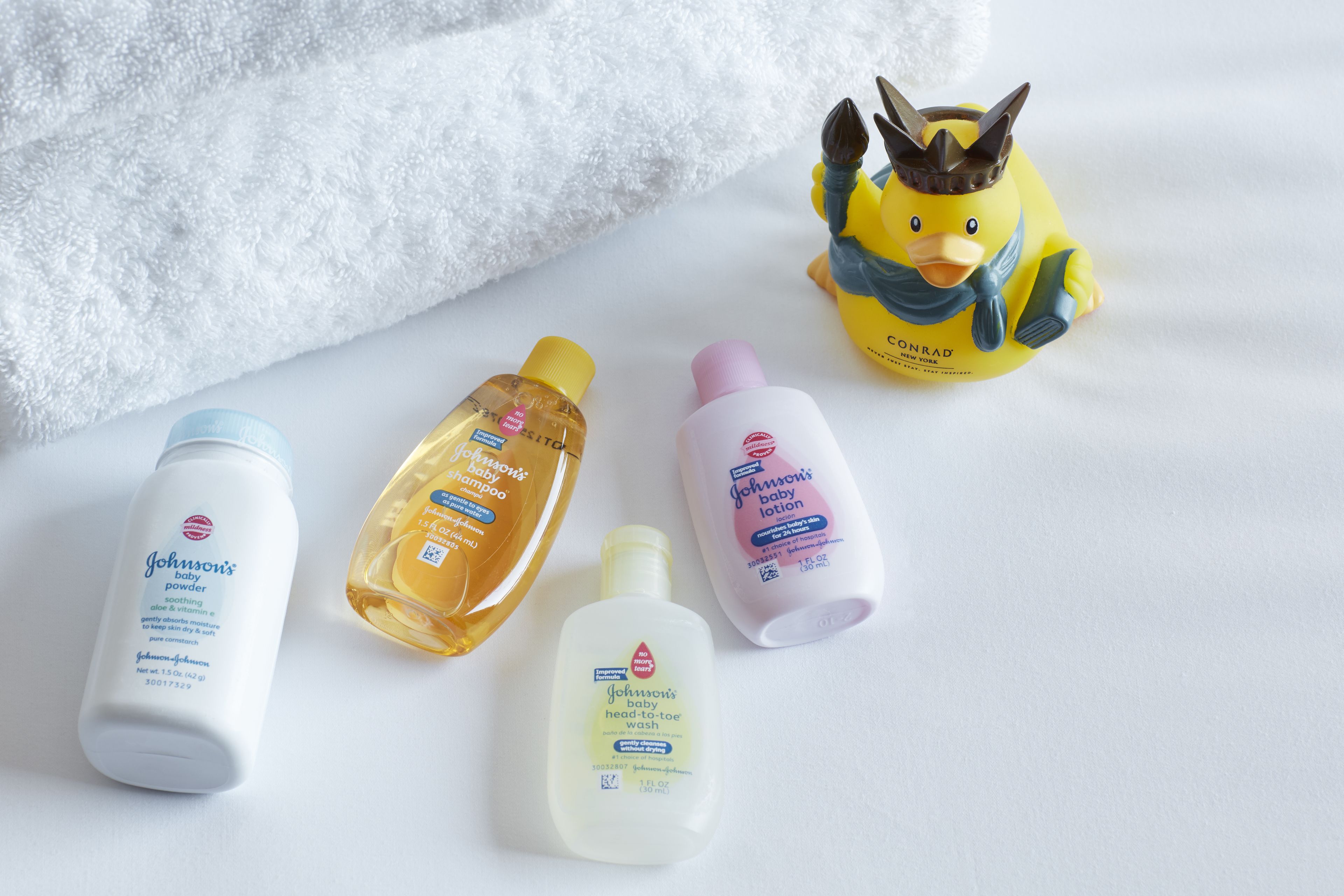 Four baby care products on a white surface with a plush white towel and a yellow duck figurine.