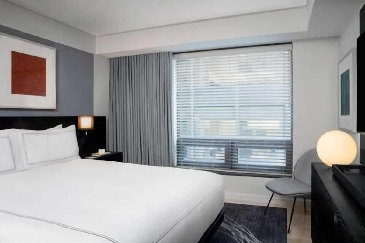 A modern hotel room with a large bed, a desk, a chair, and a window with blinds.