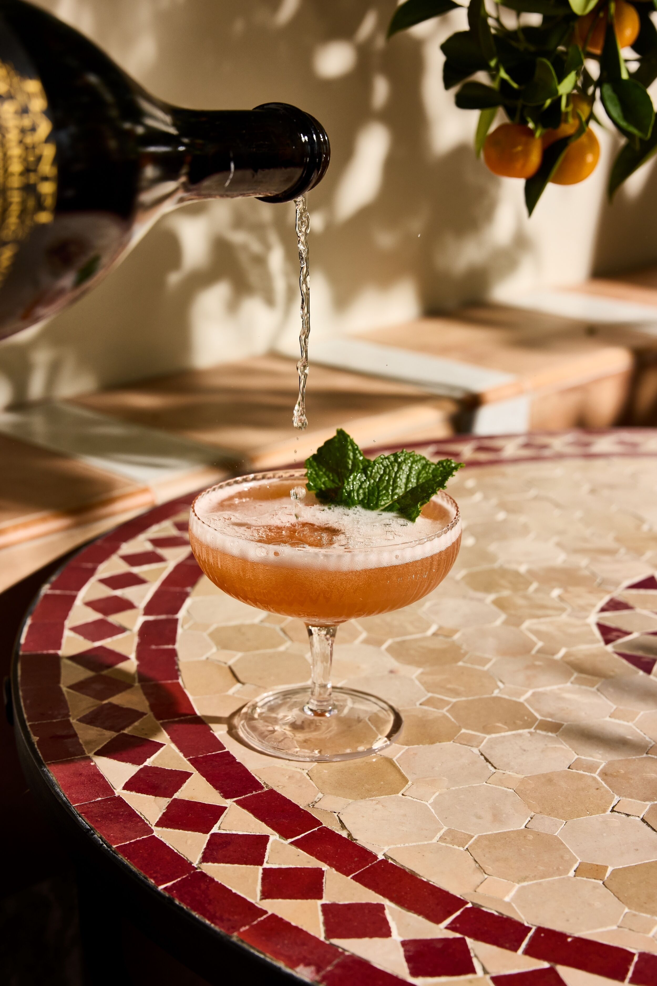 A cocktail being poured into a glass with a mint garnish, on a table with a mosaic tile pattern.
