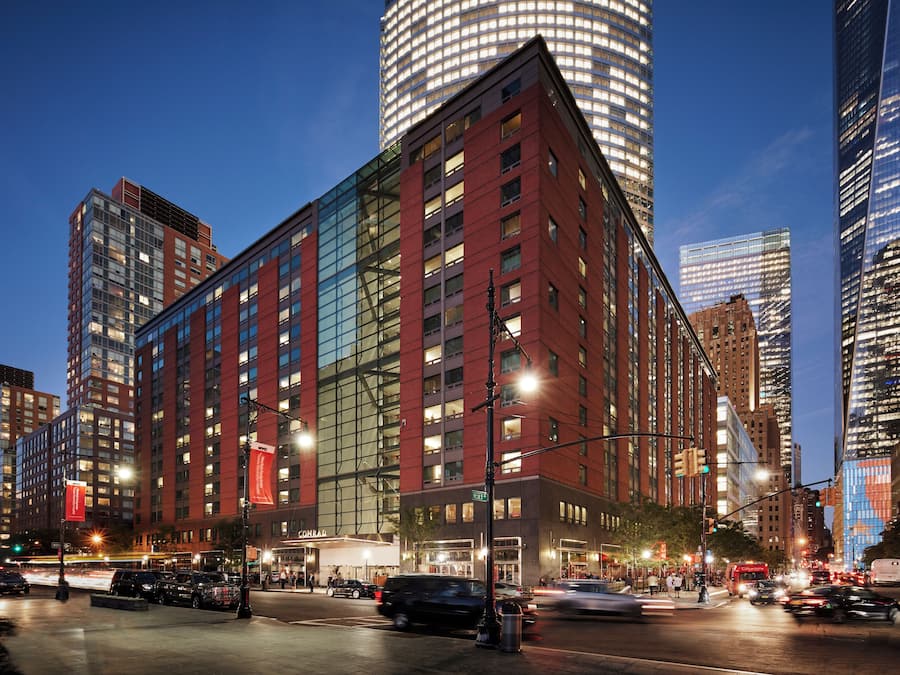Un hotel moderno con fachada de vidrio, varios pisos y una ubicación destacada en esquina dentro de un paisaje urbano.