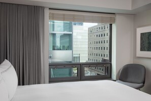 Ett hotellrum med ett stort fönster, en säng, en stol och en inramad tavla på väggen.