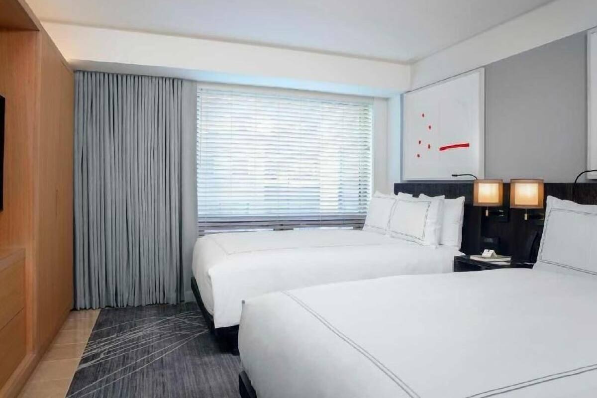 A hotel room with two beds, a large window with blinds, a wall-mounted TV, and a nightstand with lamps.