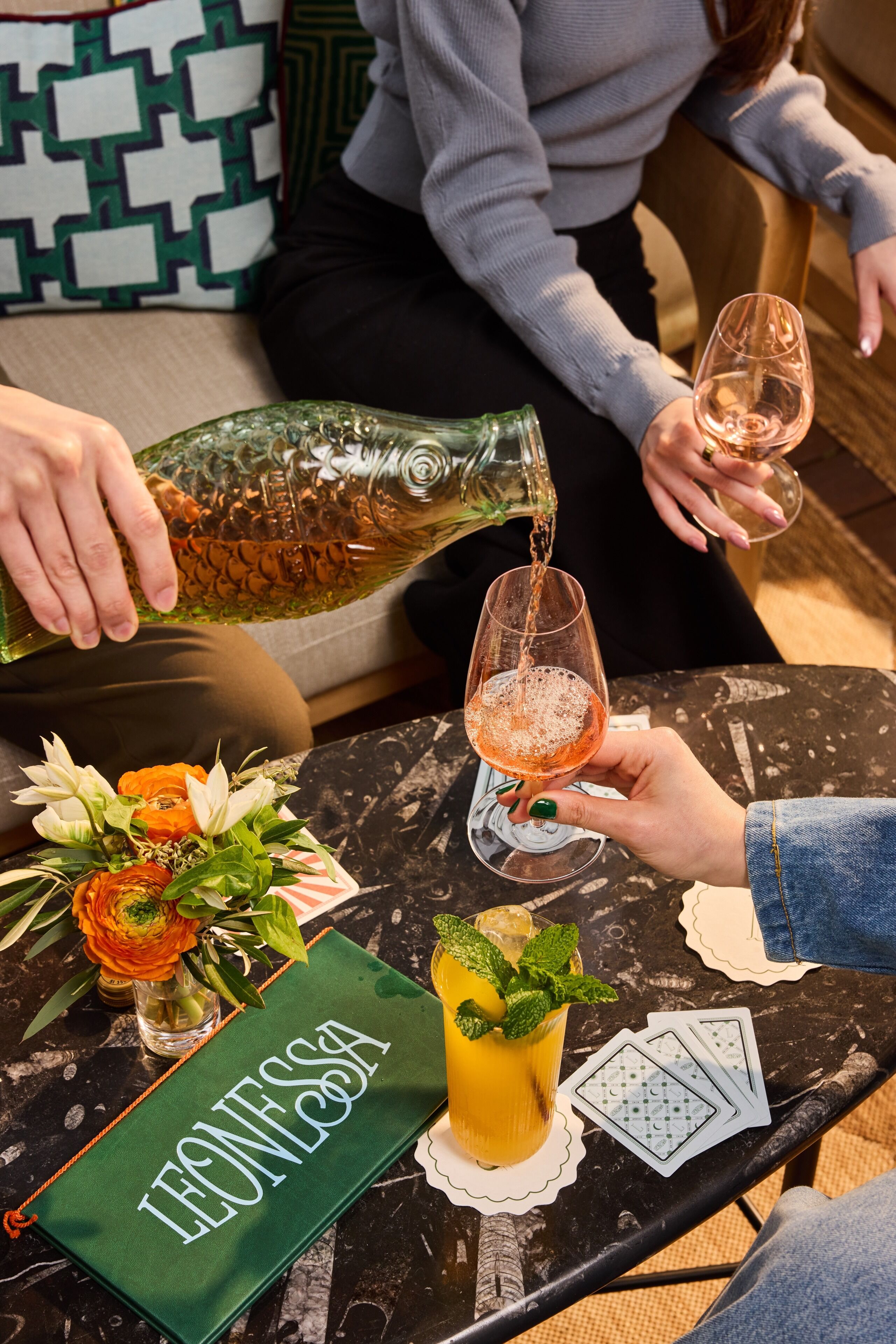 A person pouring a drink into a glass, a table with a green cloth, and a vase with flowers.