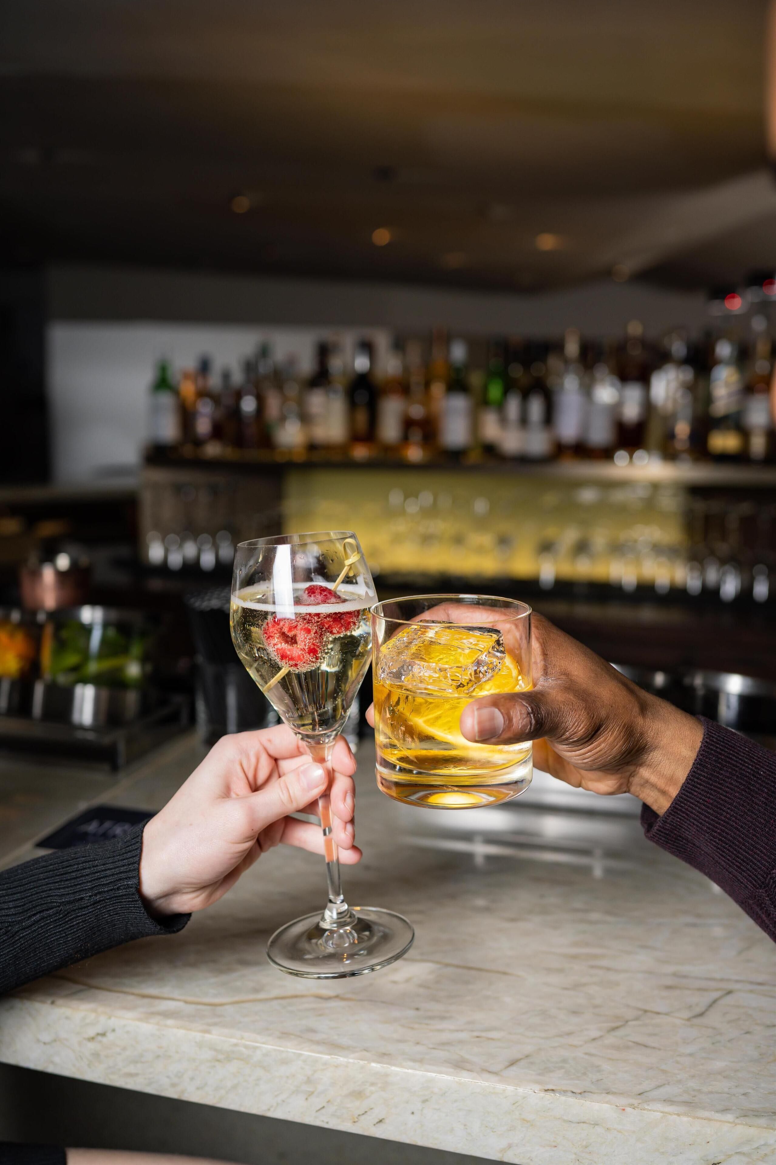 Dos manos sosteniendo vasos con bebidas en un bar, con botellas al fondo.