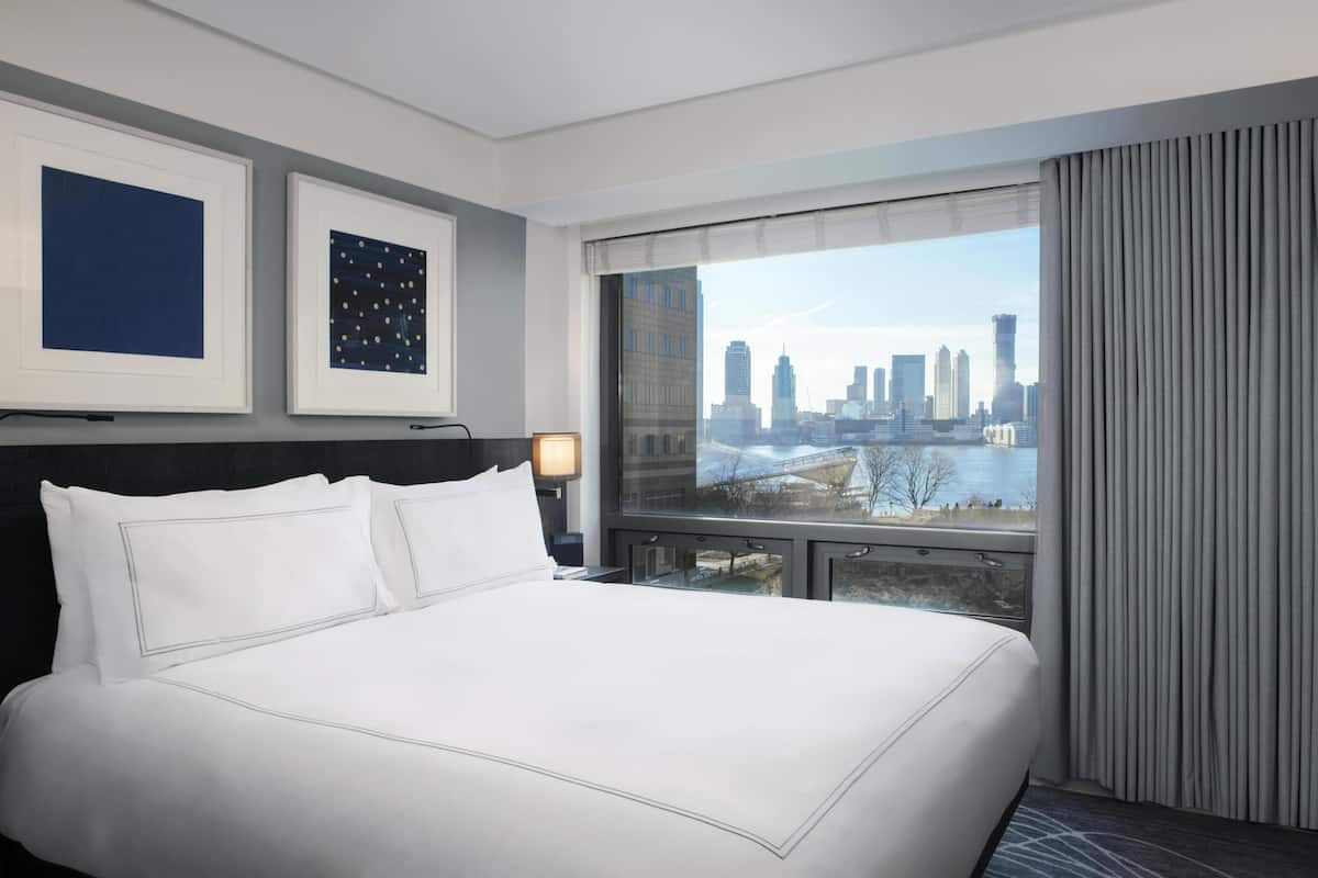 A hotel room with a large bed, a city view, a framed artwork, and a nightstand.