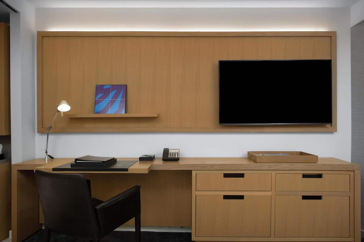 A modern office space with a wooden desk, a black chair, a mounted flat-screen TV, a desk lamp, a telephone, and a small shelf with a framed picture.