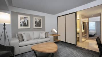 A modern hotel room with a sofa, a round wooden table, a floor lamp, a mirror, and framed artwork on the wall.