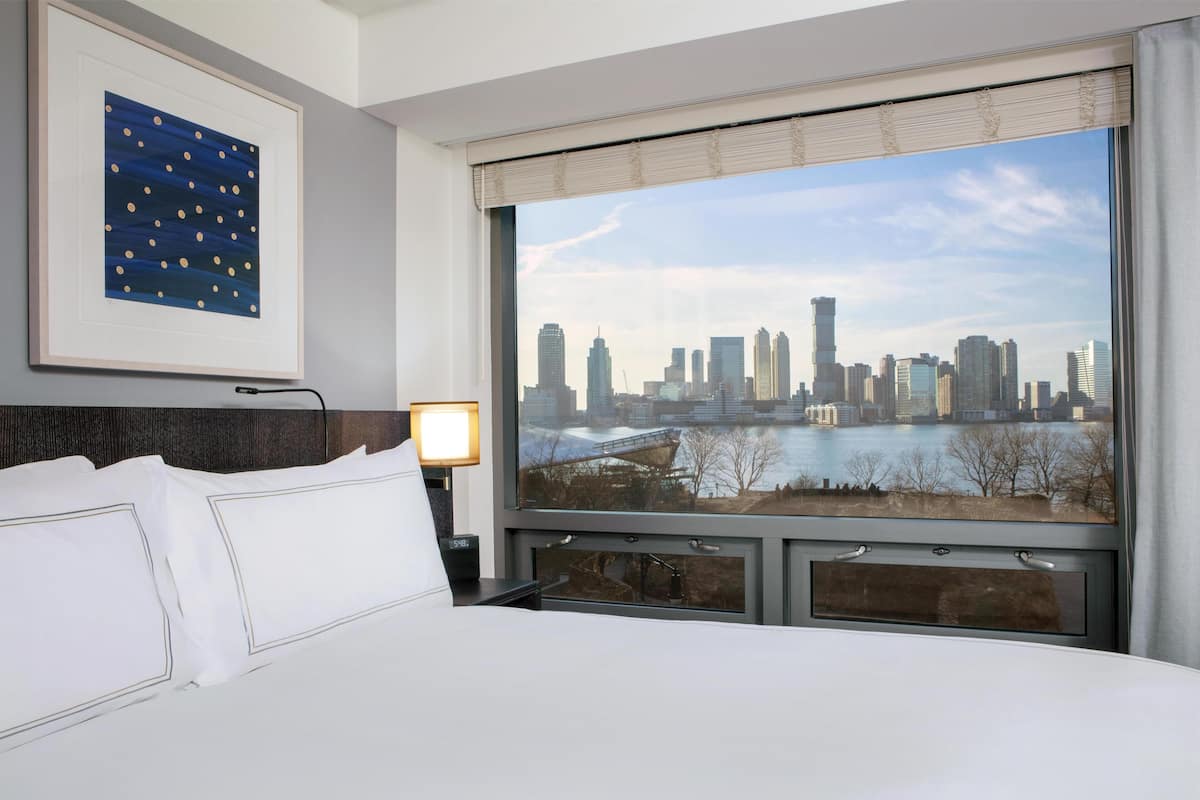 A hotel room with a bed, a framed artwork, a nightstand, a lamp, and a large window with a city skyline view.