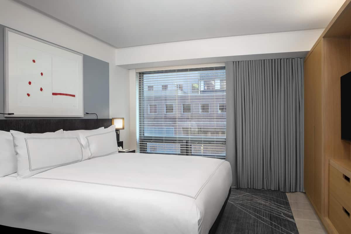 A hotel room with a large bed, a flat-screen TV, a window with blinds, and a wall-mounted artwork.