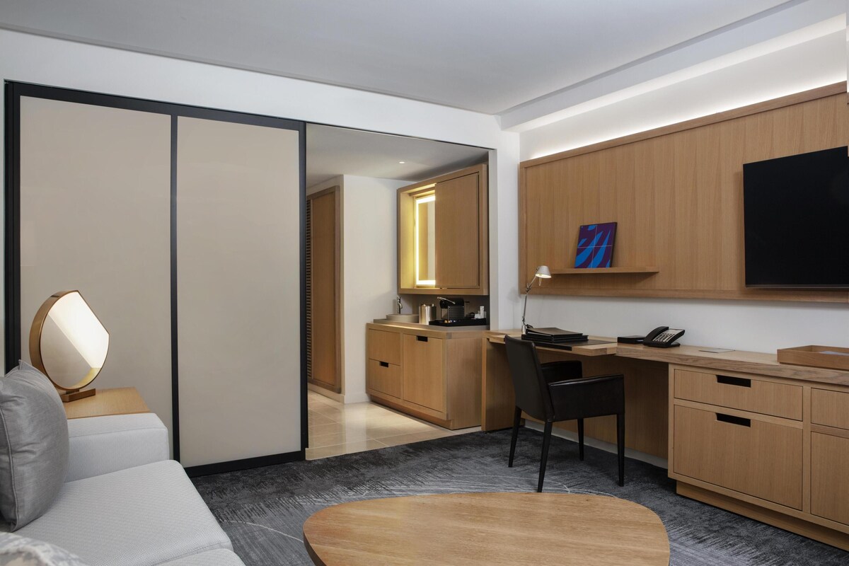A modern hotel room with a sofa, a desk, a chair, a flat-screen TV, a painting, and a sliding door leading to a bathroom.
