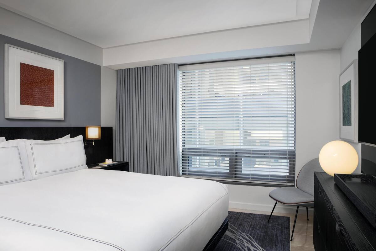 A hotel room with a large bed, a desk with a lamp, a chair, a window with blinds, and a framed picture on the wall.