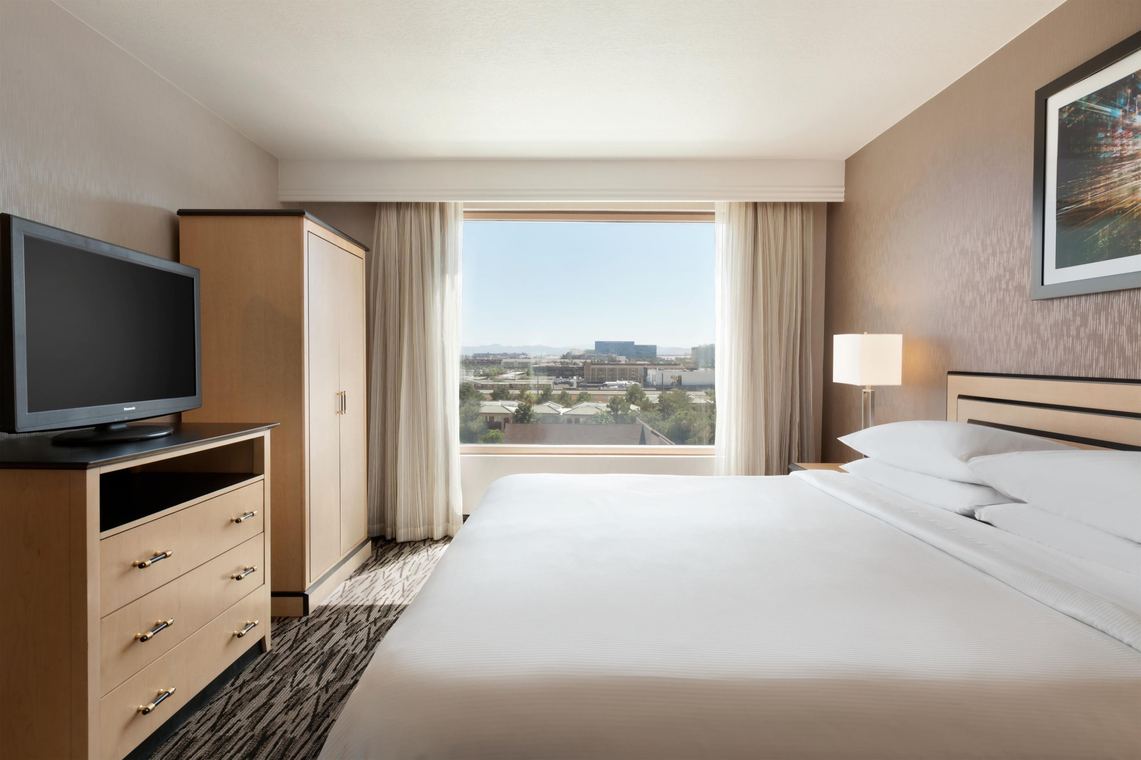 A hotel room with a bed, a television on a stand, a wardrobe, a window with curtains, and a painting on the wall.