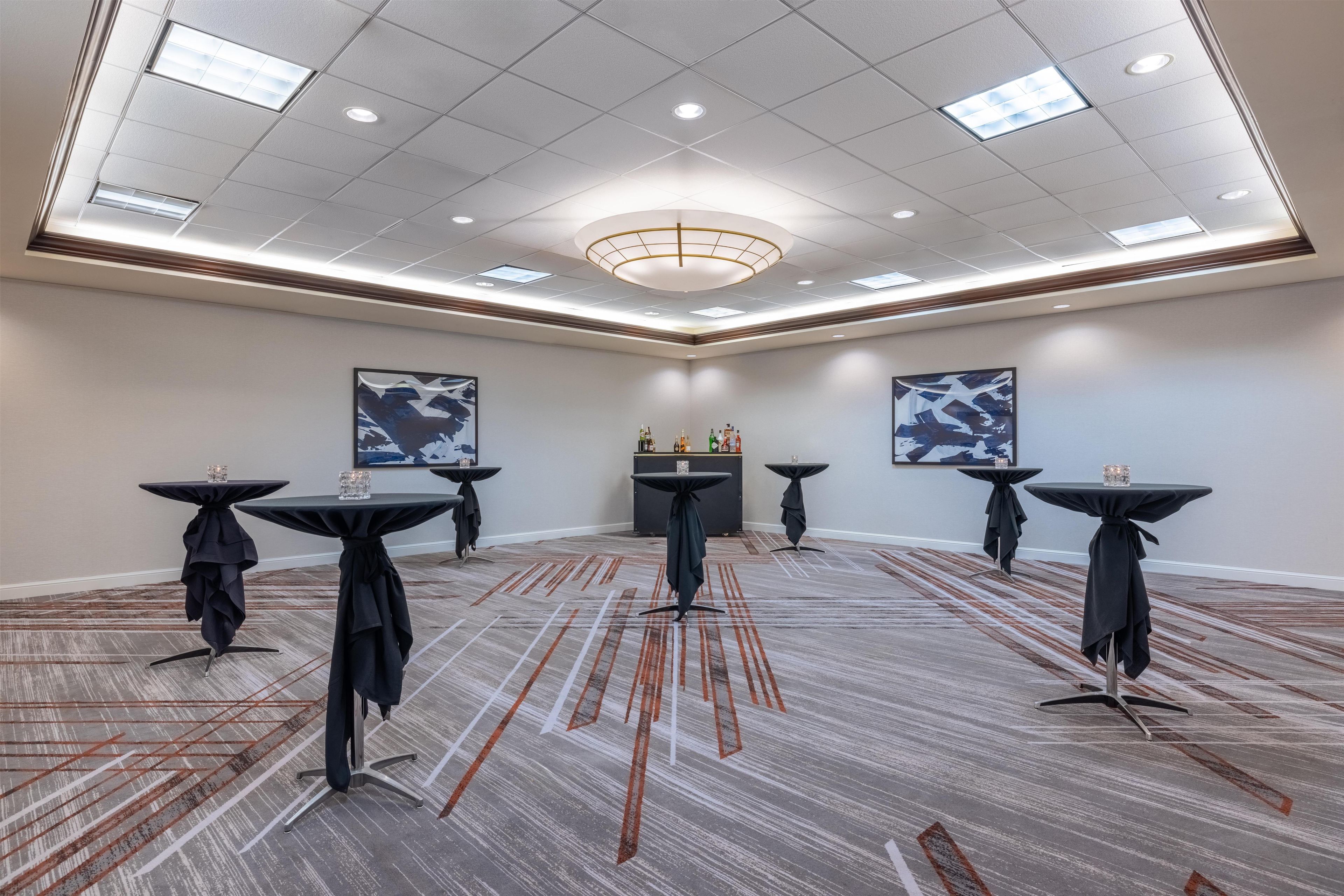Un salón de conferencias con techo alto, iluminación empotrada y un candelabro central. Seis mesas con manteles negros, cada una con un vaso encima. El salón tiene una alfombra estampada y dos grandes cuadros abstractos en las paredes.
