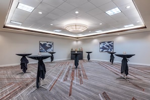 Une salle de conférence dotée d'un plafond haut, d'un éclairage encastré et d'un lustre central. On y trouve six tables recouvertes de nappes noires, chacune portant un verre. La salle est agrémentée d'un tapis à motifs et de deux grandes peintures abstraites accrochées aux murs.